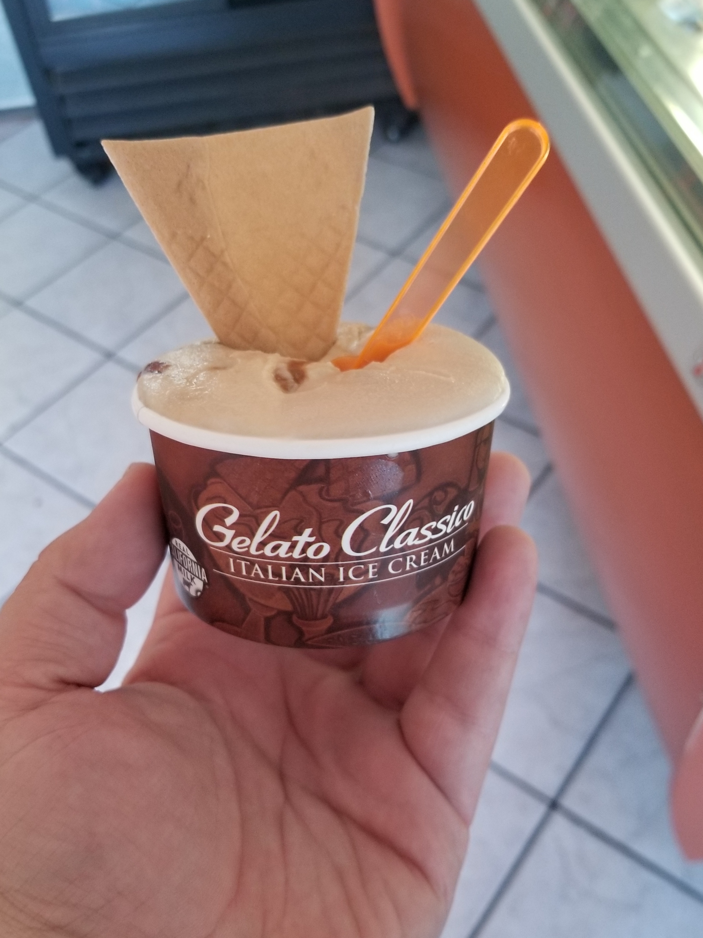 A hand holding a cup of gelato with a triangular wafer cookie and orange plastic spoon.