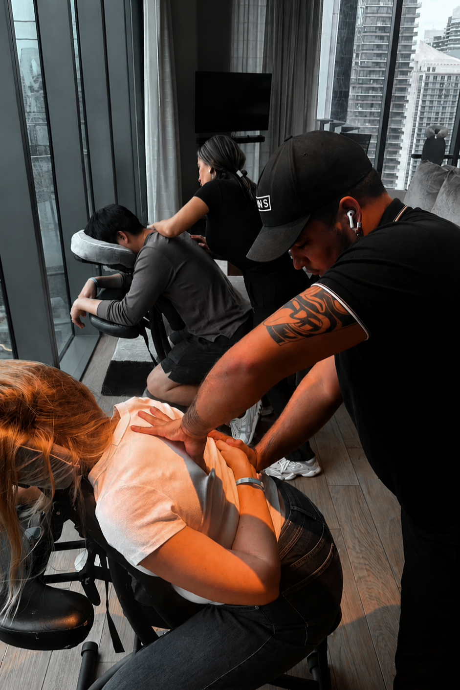 A woman receiving a chiropractic adjustment from a male chiropractor while a woman in the background is getting a massage by another practitioner in a high-rise building.