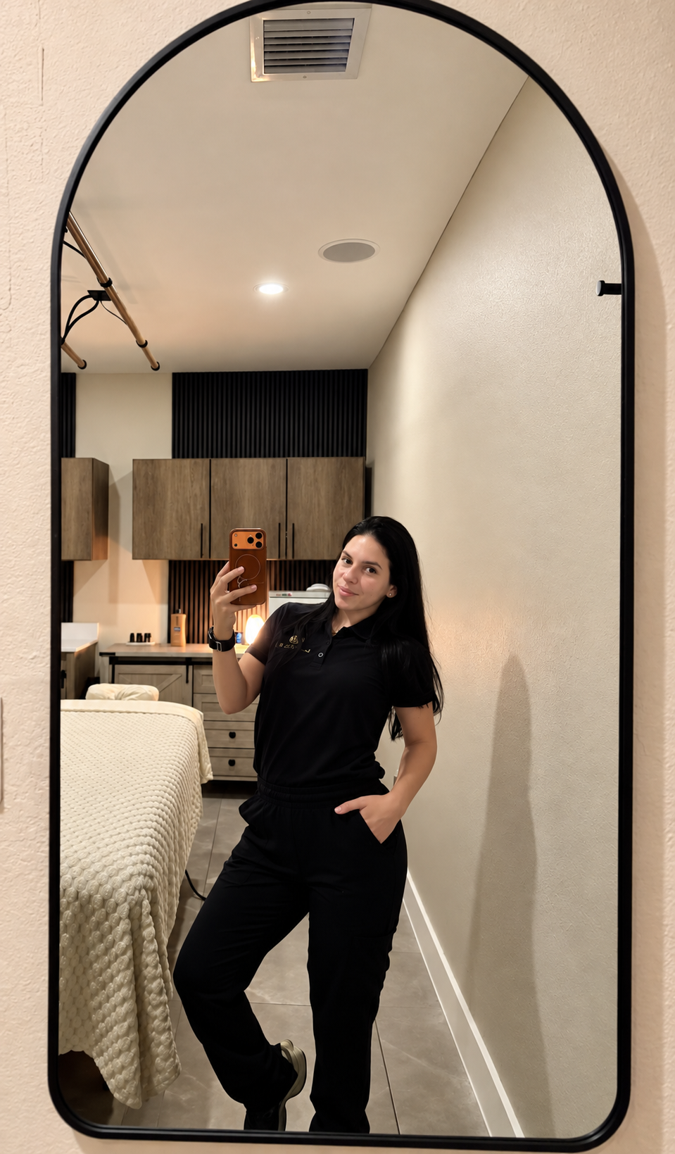 A woman taking a selfie in a large mirror in a room with beige walls, bedroom furniture, and a bed with a textured cream-colored blanket.