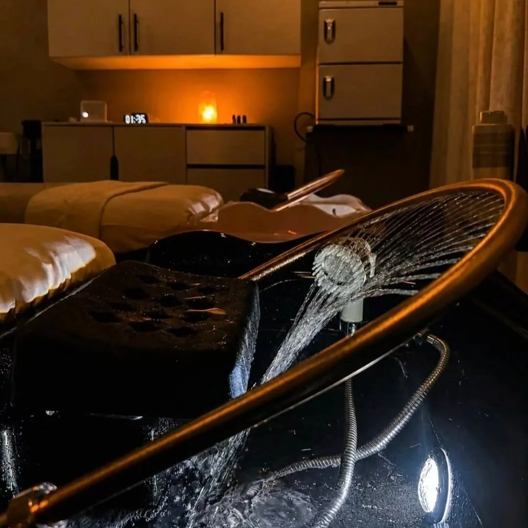 A glass shower head sprays water into a bathroom, with a bedroom visible in the background featuring beds, a clock, and cabinets.