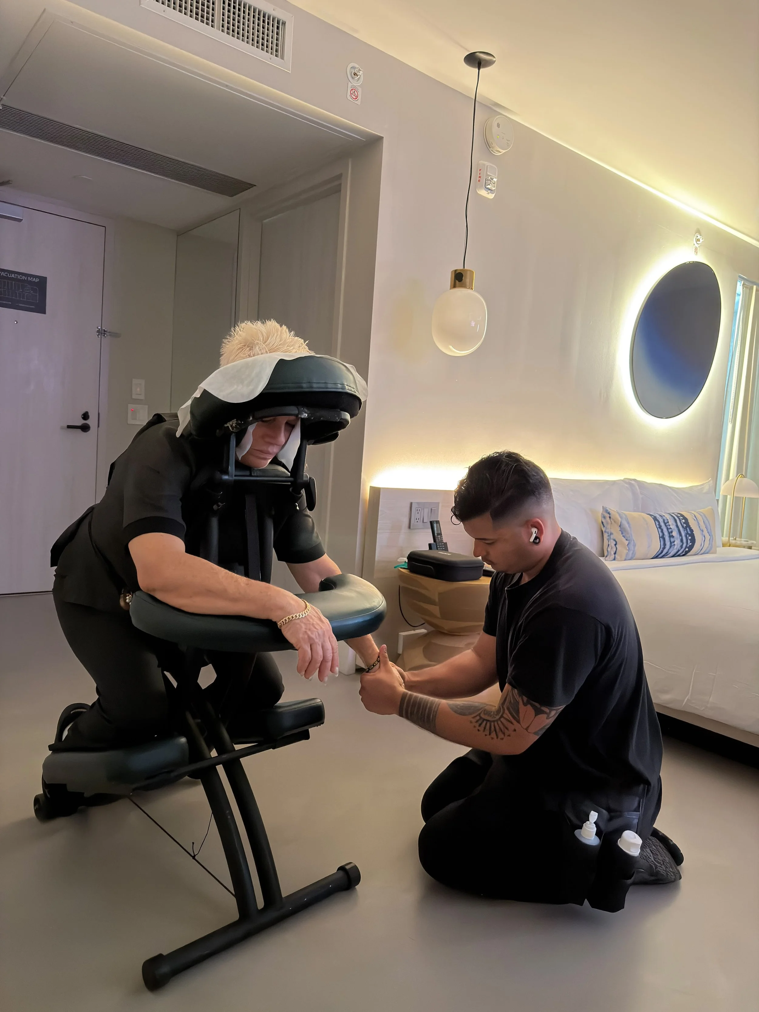 A man with headphones [AirPods] kneeling on the floor and holding the hand of an elderly woman who is sitting on an exercise bicycle. The woman is dressed in black with a light-colored scarf on her head. The room has modern lighting, a bed with pillo