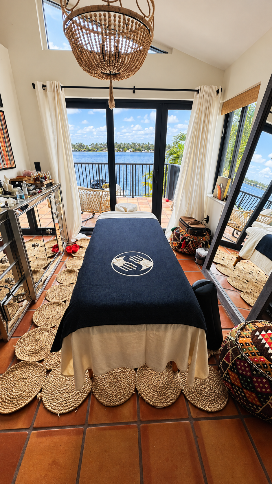 A massage table with a dark blue cover and a white skirt, situated indoors with a view of a lake through large glass sliding doors. The room has terracotta tile floors, woven circular rugs, and white curtains. Outside, there is a small balcony with a
