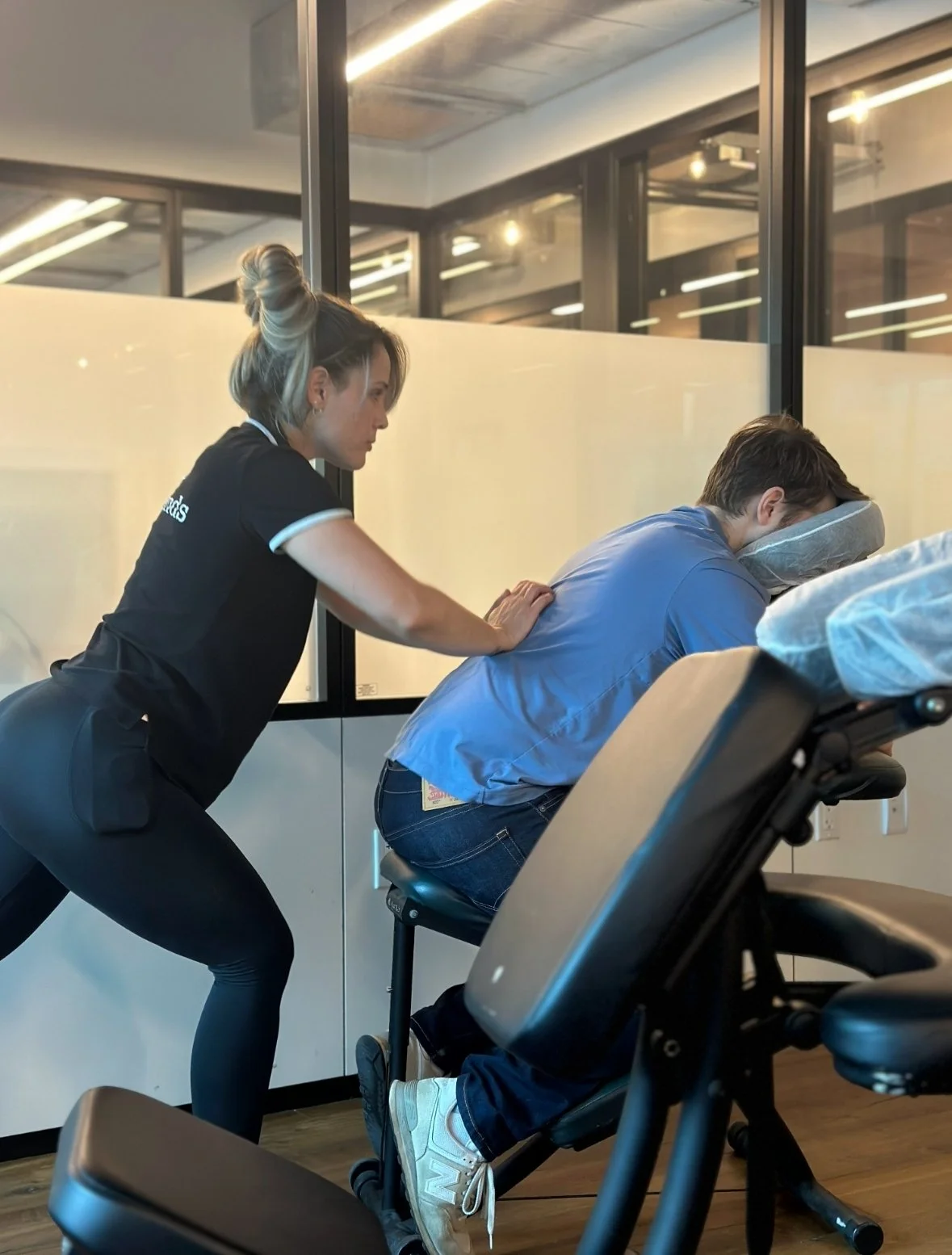 A woman massage therapist providing a back massage to a man seated in a massage chair inside a modern room with glass walls and ceiling lights.