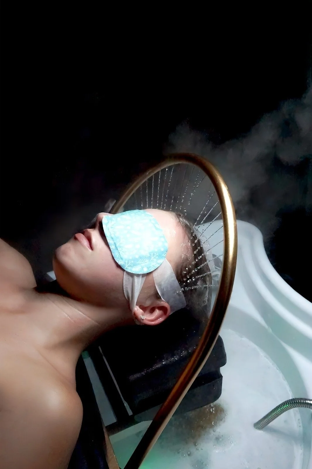 A woman is receiving a steam facial treatment while lying down with a sleep mask on her eyes, in a spa or wellness setting.