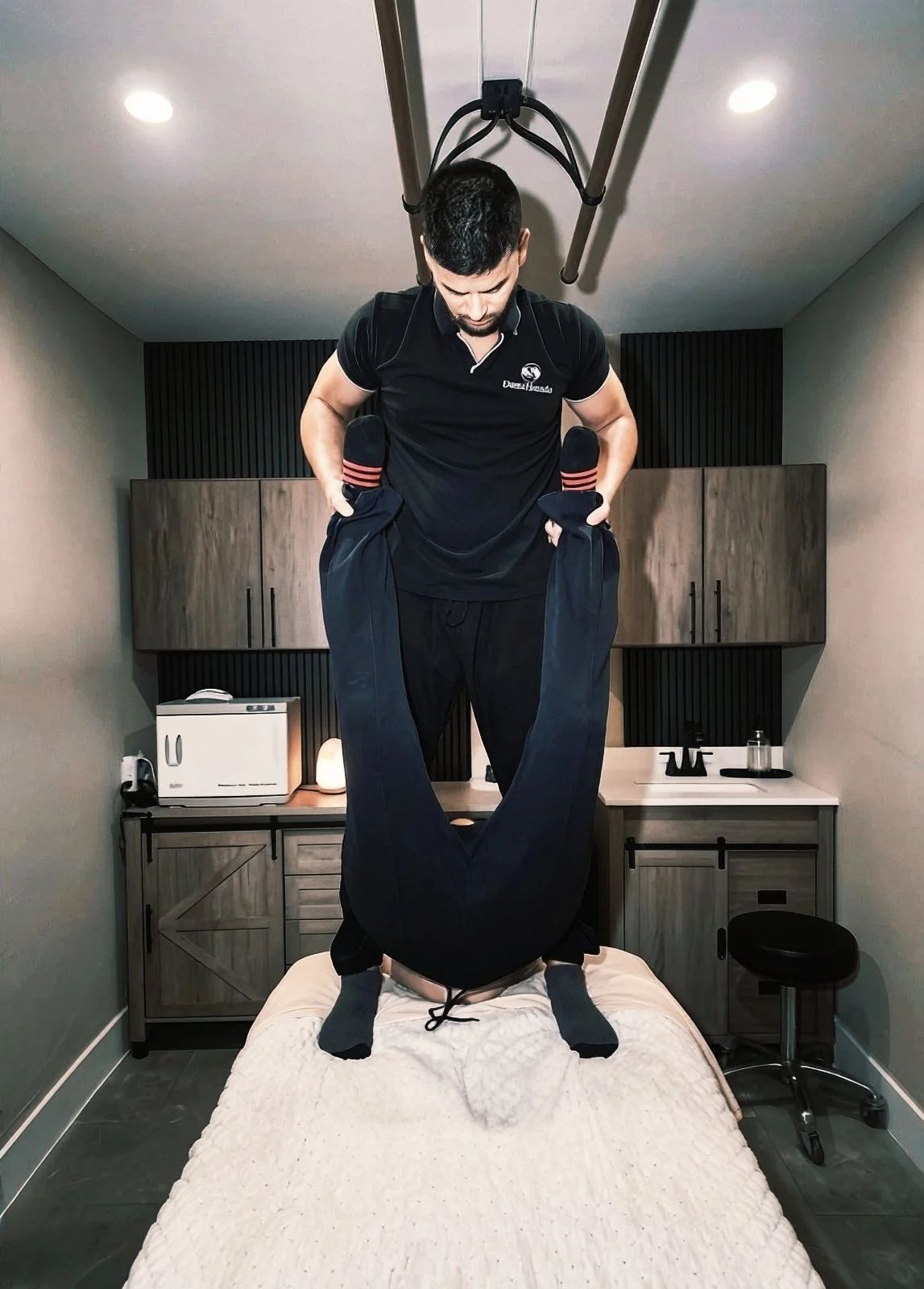 A man is standing on a massage or therapy table in a small room, holding another person upside down by their ankles, with the person’s head and upper body hanging down. The room has wooden cabinets, a small lamp, a toaster, a soap dispenser, and a st