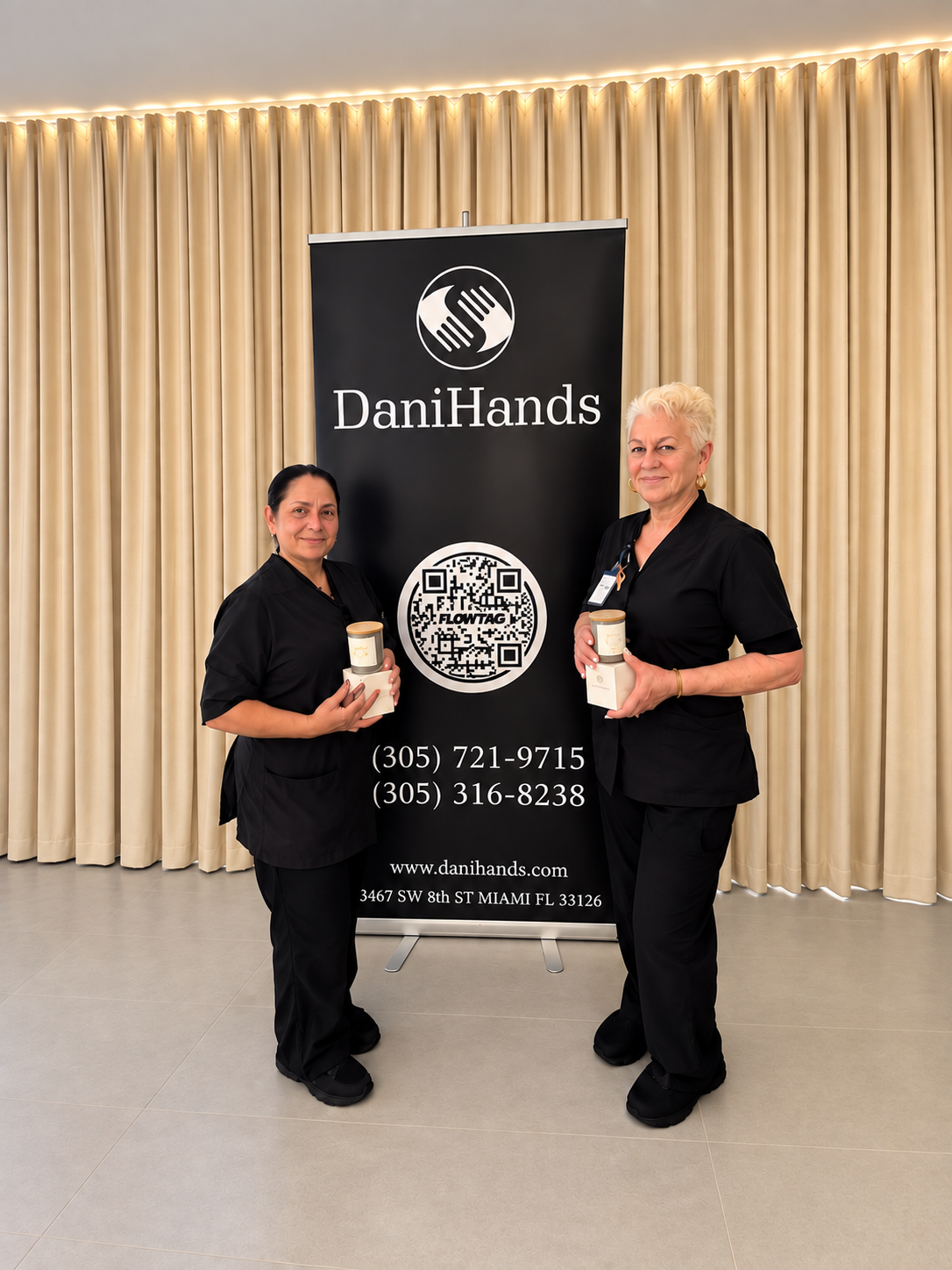 Two women dressed in black uniforms standing in front of a Danihands banner, holding candles or jars with a label, at an event in an indoor venue with beige curtains.
