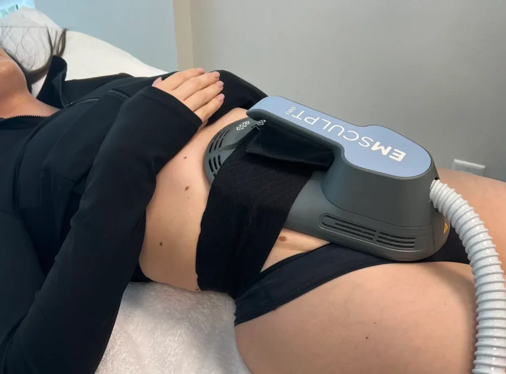 Person lying on a medical bed with a machine labeled 'EMSculpt Neo' on their abdomen, undergoing a non-invasive body contouring procedure.