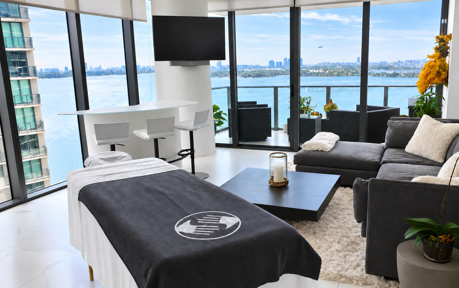 Modern apartment living room with a view of the city skyline and river, containing a gray sofa with pillows, a black coffee table with a candle, a bed with a black cover, a white curved bar with stools, and a balcony with outdoor seating and potted p