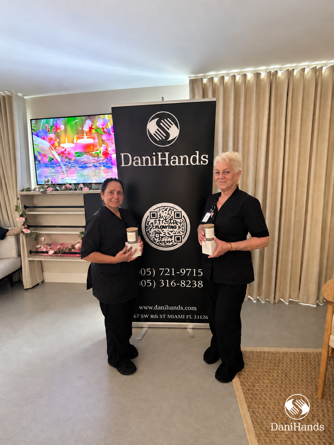Two women in black uniforms standing in front of a DaniHands banner, each holding a jar of product, at an indoor event with curtains and a colorful digital screen in the background.