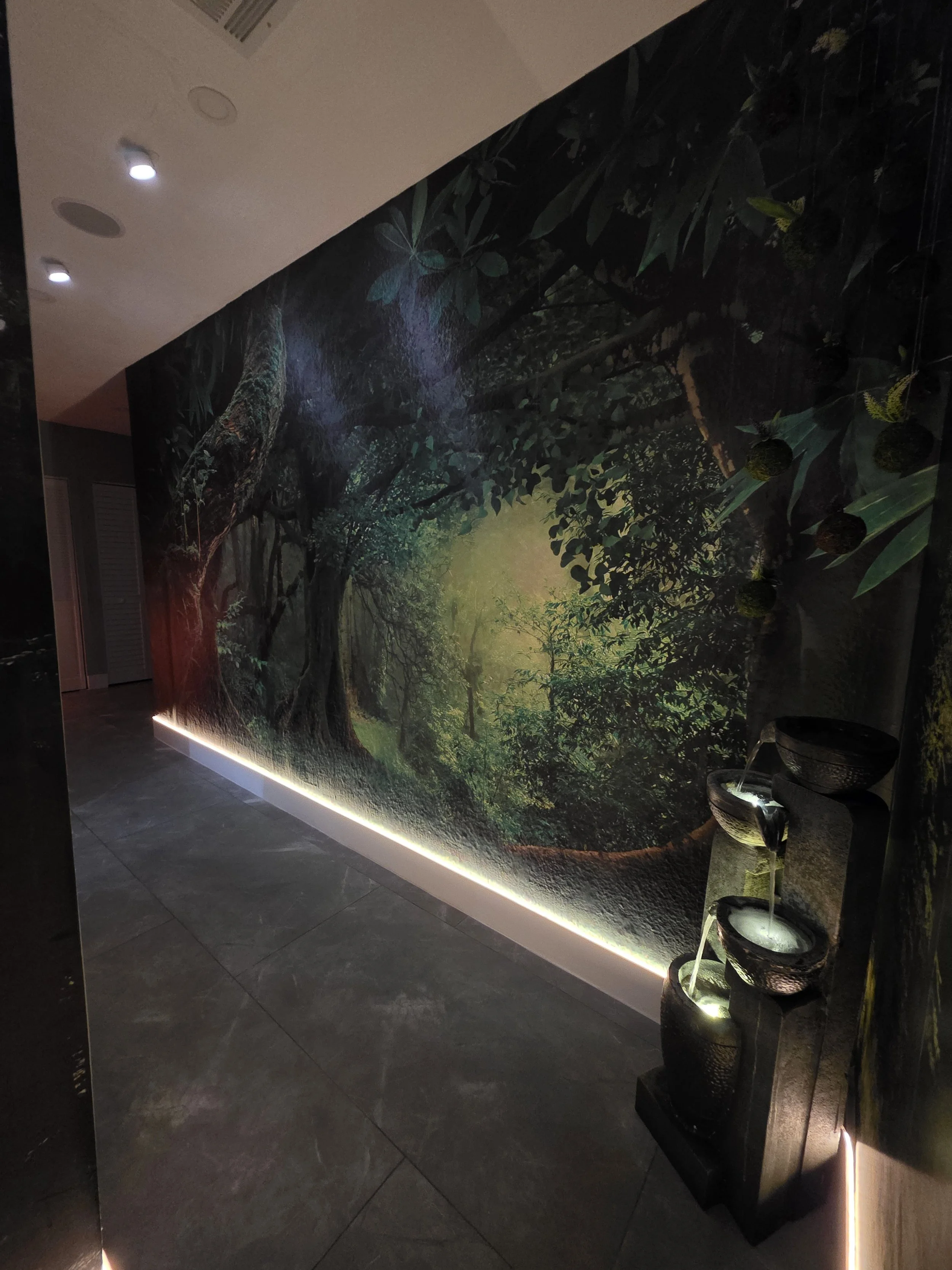 A hallway wall mural depicting a forest with trees and green foliage, illuminated by subtle lighting along the bottom edge. To the right is a modern fountain with a black textured finish, featuring multiple tiers with water flowing from the top.
