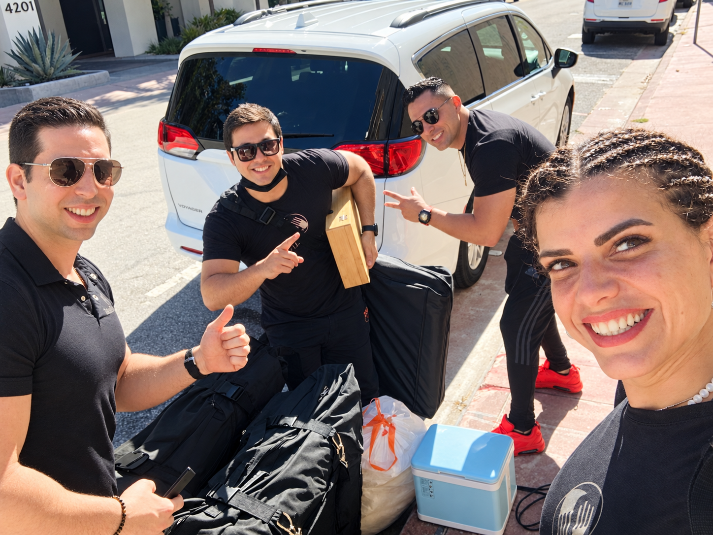 Group of five young adults smiling and taking a selfie on sidewalk near parked car, carrying luggage for travel.