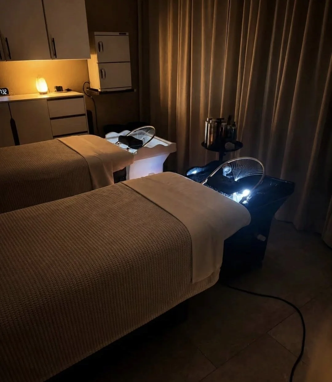 A dimly lit spa treatment room with two massage beds, each covered with a beige blanket, and small lamps providing soft lighting. There are bedside tables with spa tools, and a cabinet with a clock showing 7:32. Curtains cover the windows, and the overall atmosphere is calm and relaxing.