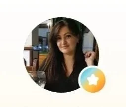 A woman with long brown hair smiling, indoors with a computer monitor in the background, circular profile picture.