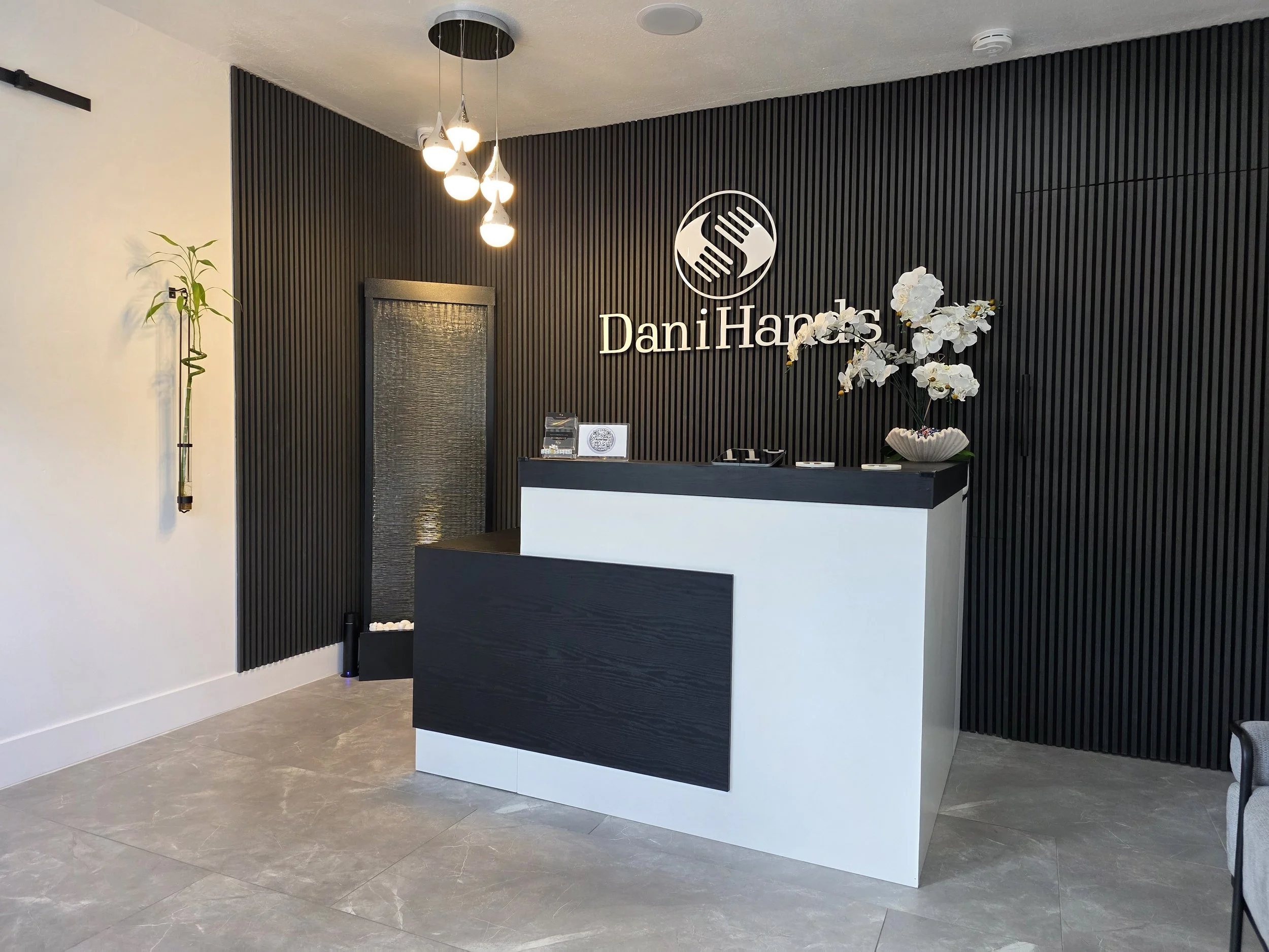 Modern office reception area with black slatted accent wall, white reception desk, decorative white orchids, black door, and hanging light fixture.