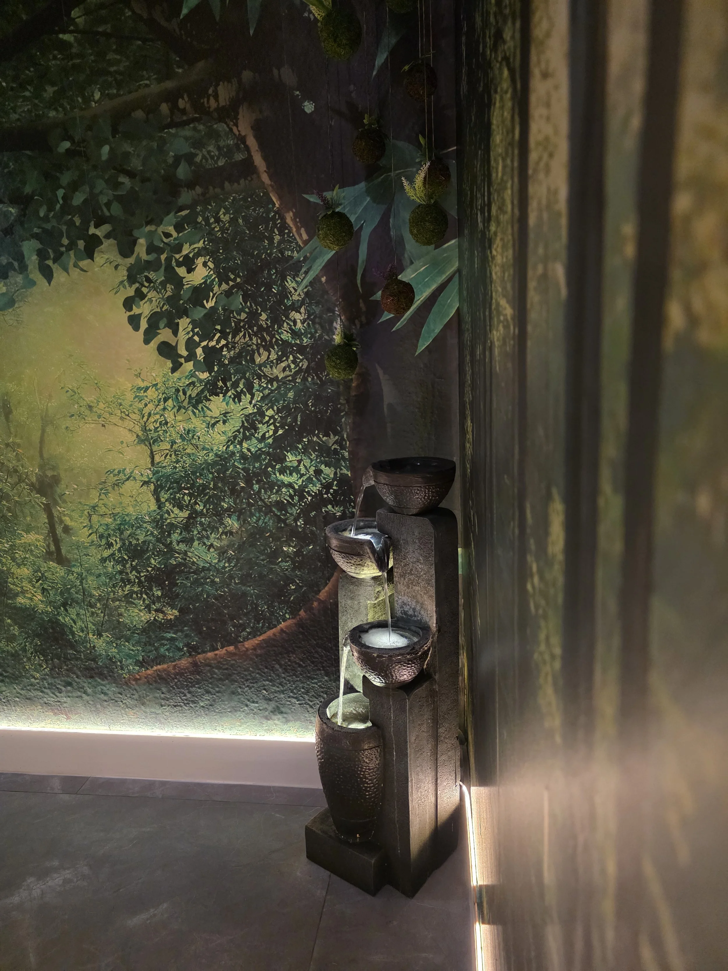 Indoor water fountain with flowing water, set against a backdrop of a nature-themed mural featuring trees, foliage, and large leaves.