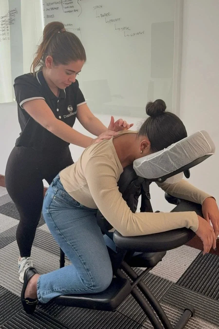 A woman receiving a massage from a therapist at a wellness or physical therapy clinic, with a whiteboard and writing visible in the background.