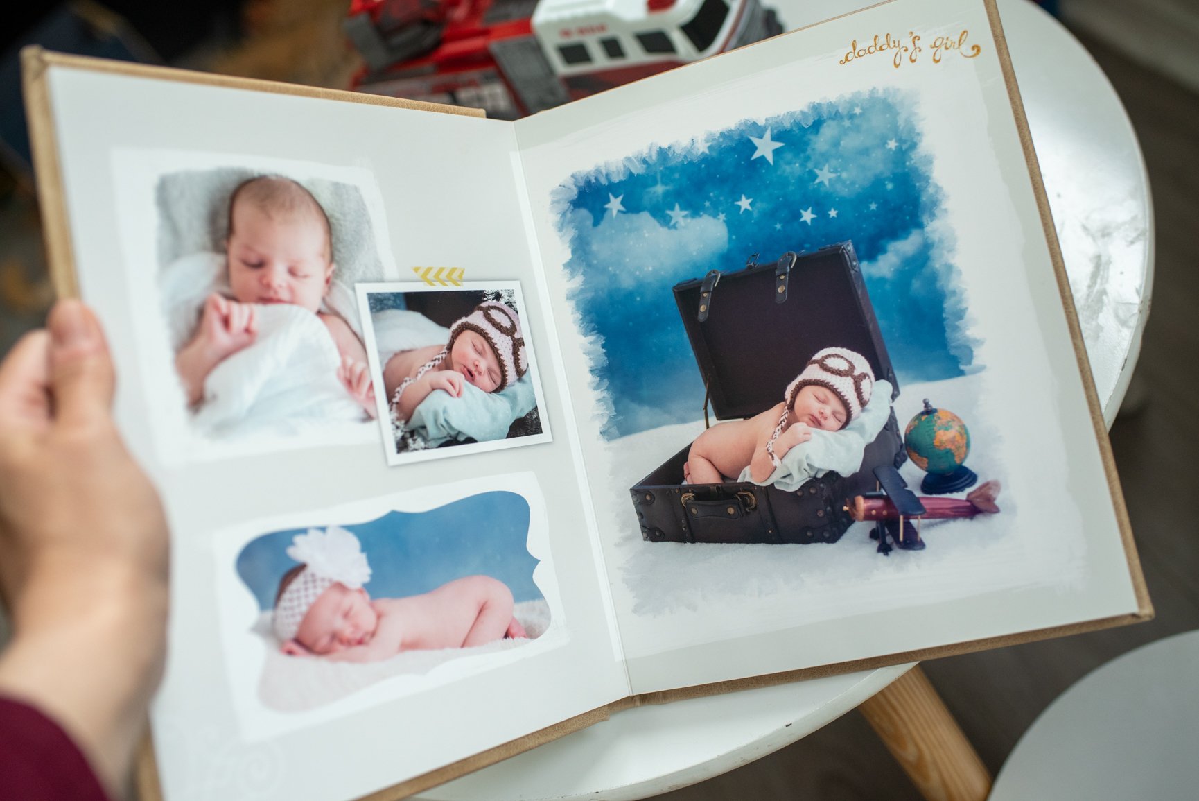 Open baby photo album with pictures of a sleeping newborn girl, including a large picture of the girl lying in an open suitcase with a travel theme backdrop, a globe, a small airplane toy, and a rock.