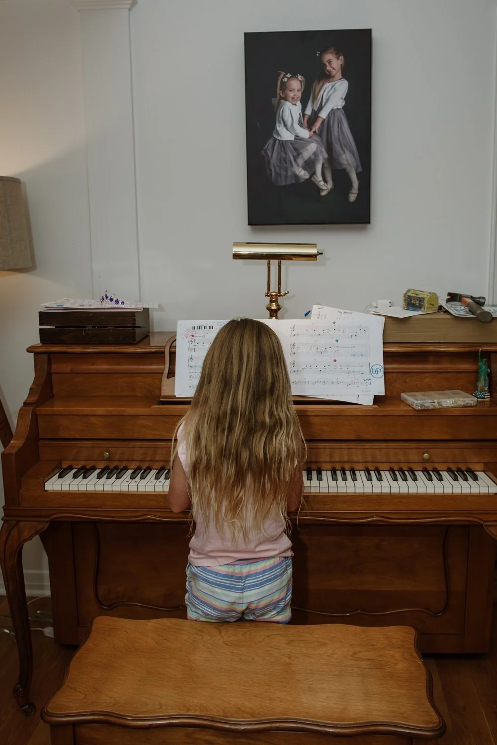 A young girl with long, wavy blonde hair sitting at an upright wooden piano, looking at sheet music. There is a photograph of two girls, one younger and one older, hanging on the wall above the piano. The room has various items including a table lamp