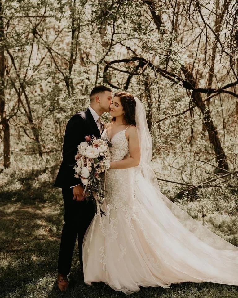 Bride and groom standing close in a forest, bride holding a bouquet of flowers, groom kissing bride's temple, wedding attire with bride in a lace dress and veil, groom in a black suit, trees in background