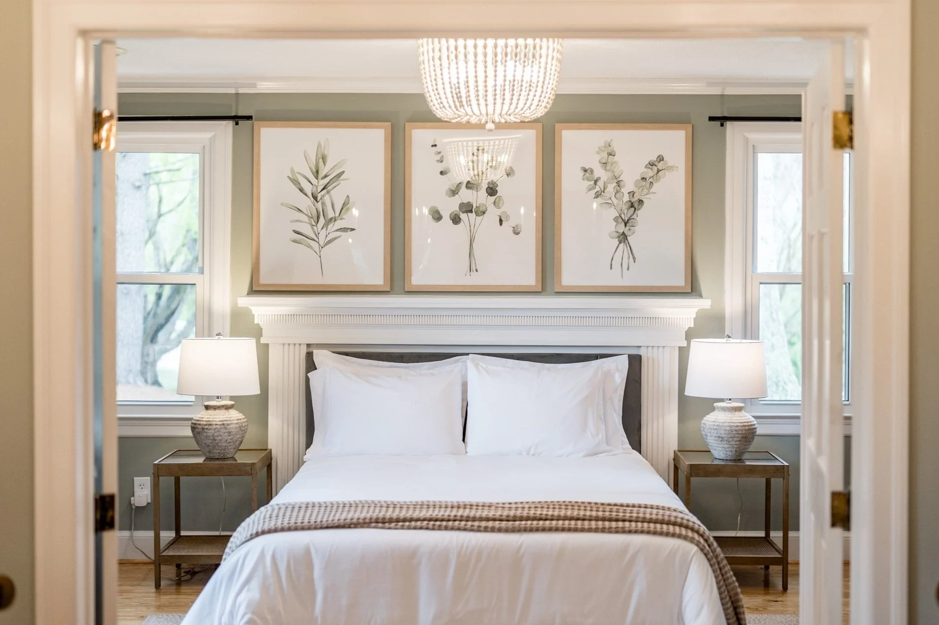 Bedroom with white bed, framed botanical art on green wall, two windows, two matching nightstands with ceramic lamps, chandelier reflected in mirror