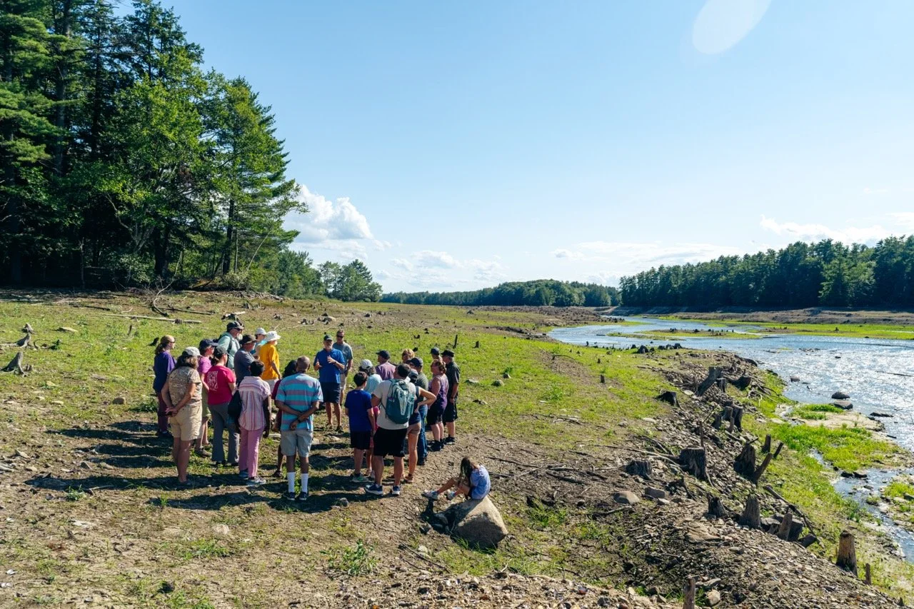 A group of ~15 people gather around Michael Shaughnessy, who is giving a talk on a grassy open space next to the Presumpscot River.