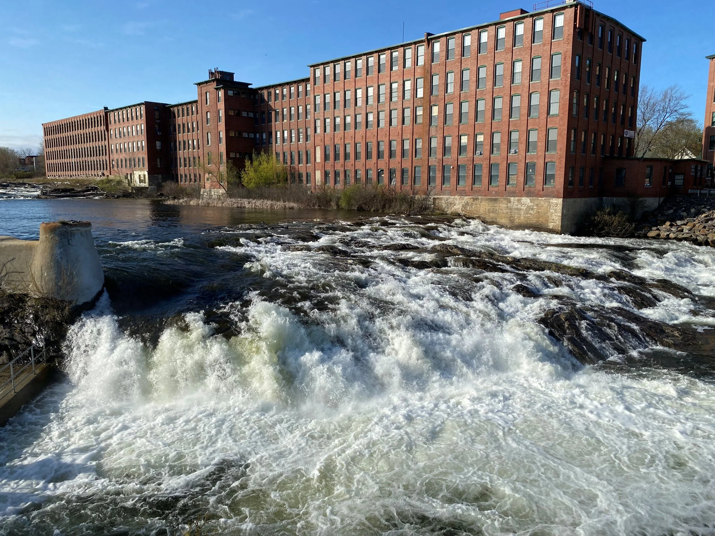Dana Warp Mill, a large five-story brick building with a full grid of windows on every face. The mill is located on the Presumpscot Riverfront, above Saccarappa Falls