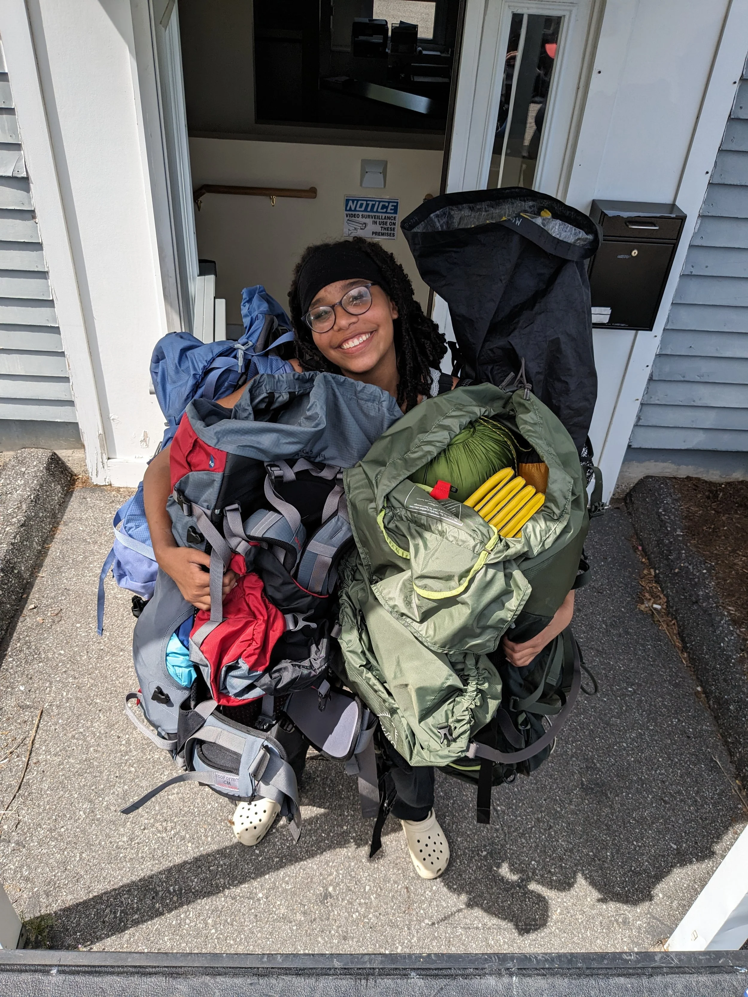 A woman smiles while holding several large bags of outdoor gear