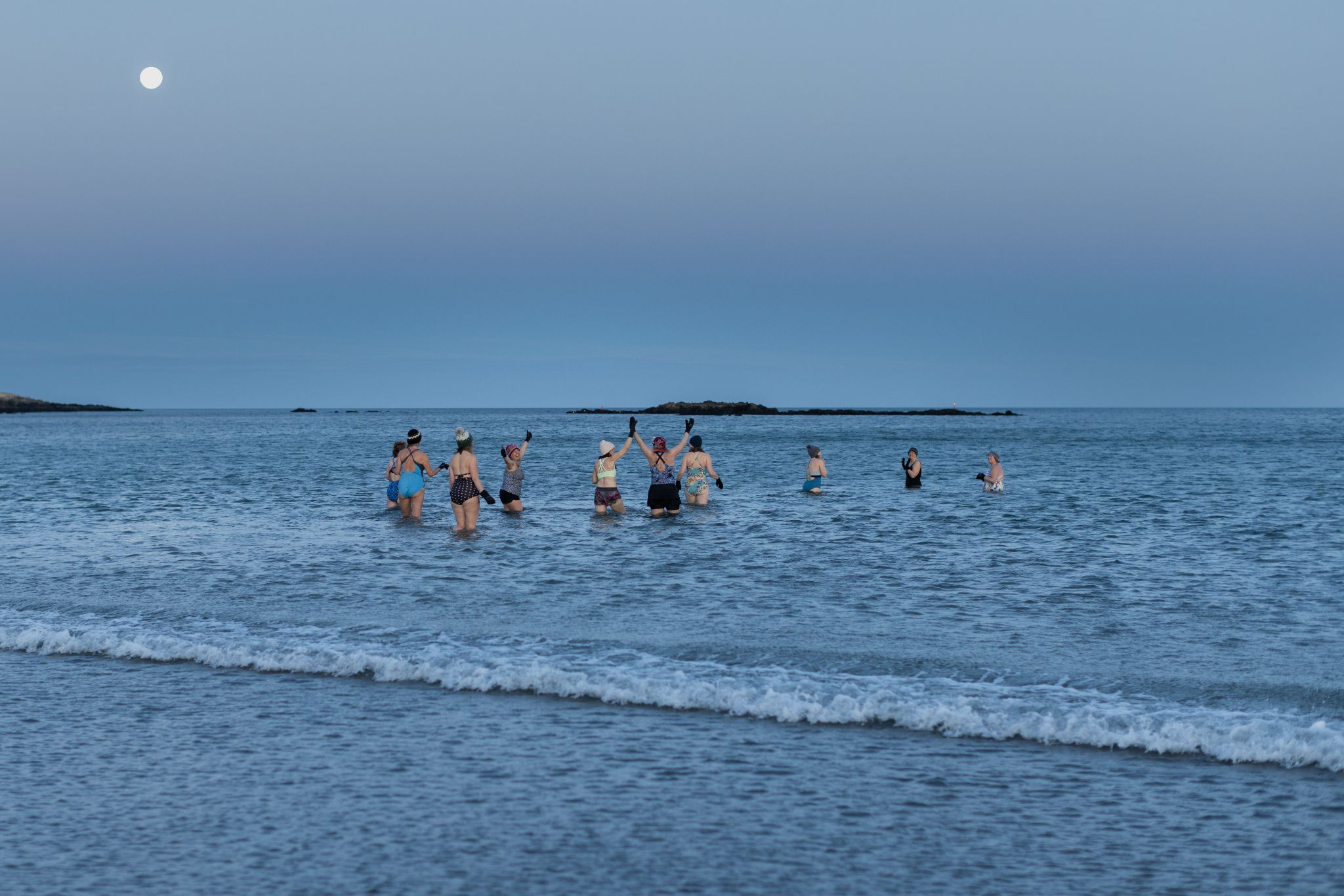 A group of around 10 women wear bathing suits and winter hats while they stand in the ocean under a full moon