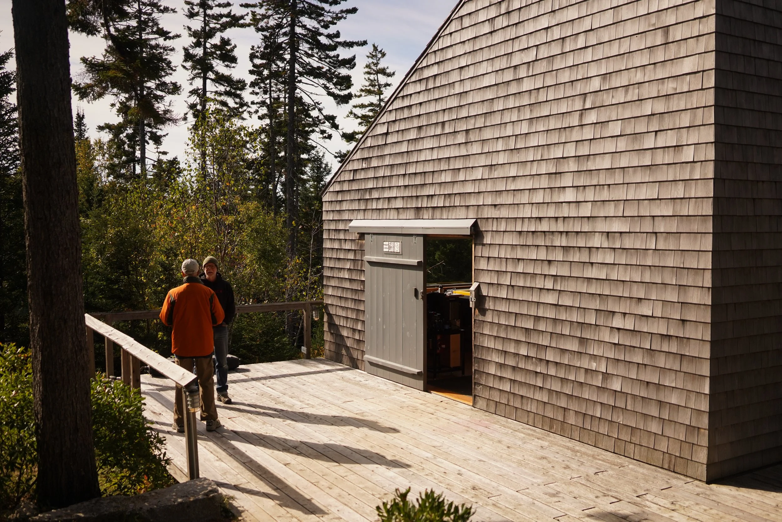 Exterior view of Haystack Mountain School building with cedar siding and 2 people standing in the distance looking at trees