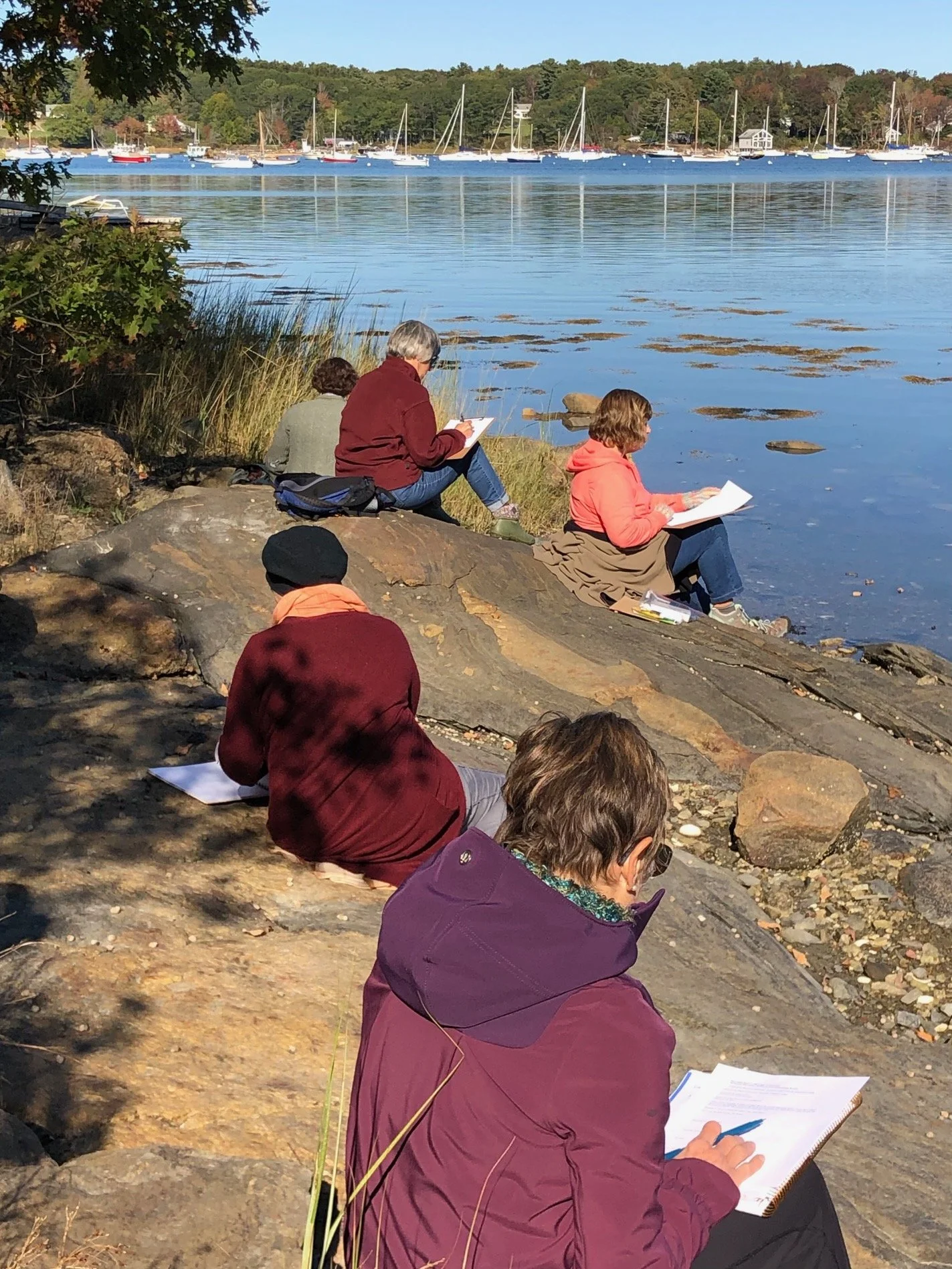 Five women sit on a rocky shore. The women are spread out and each hold a stack of paper and a pen.