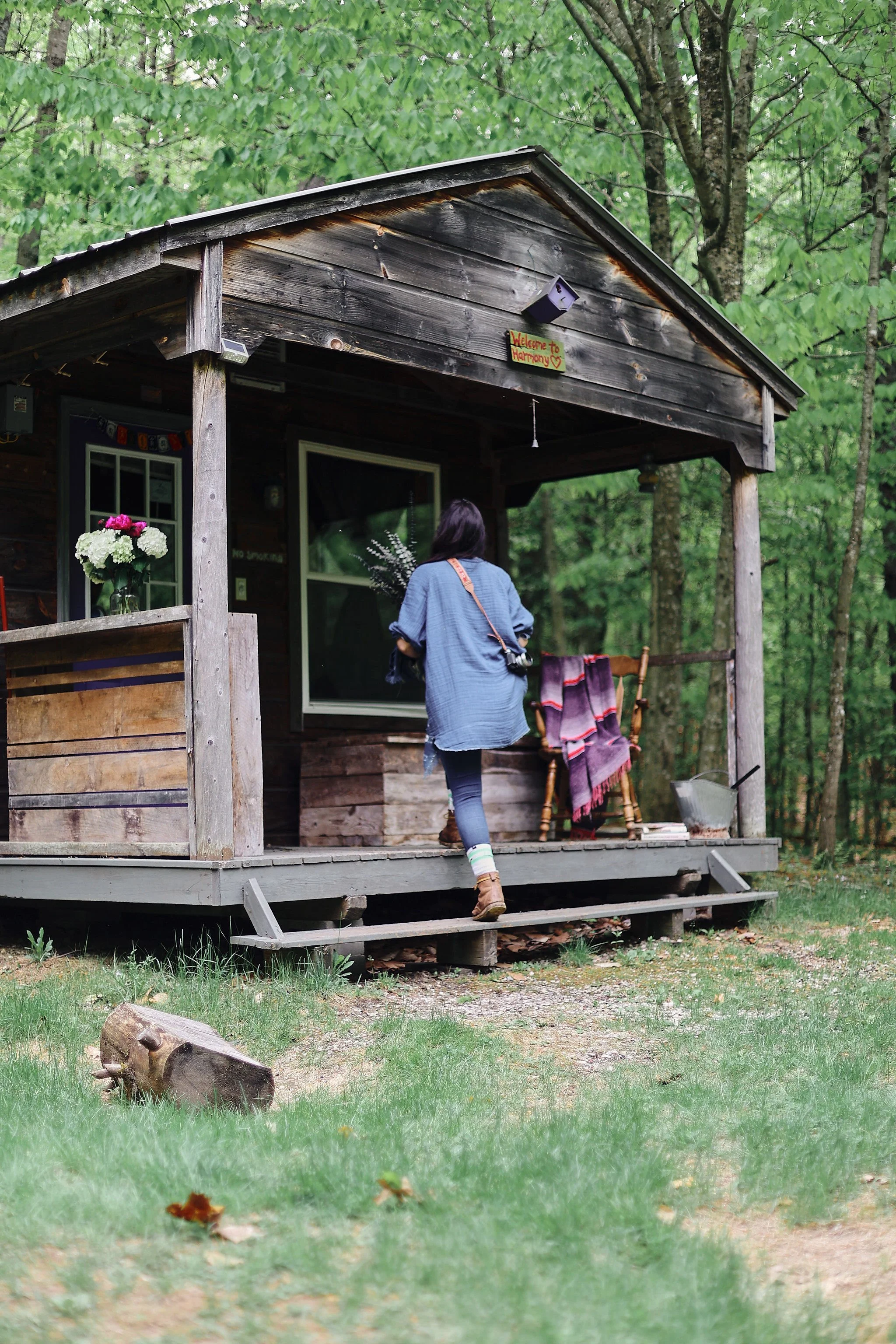 A woman, as seen from behind, walks up to the porch of a rustic cabin while holding a bouquet