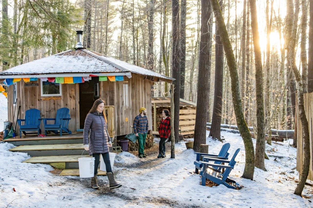 A woman in a long winter coat carries buckets away from a wooden sauna in the woods