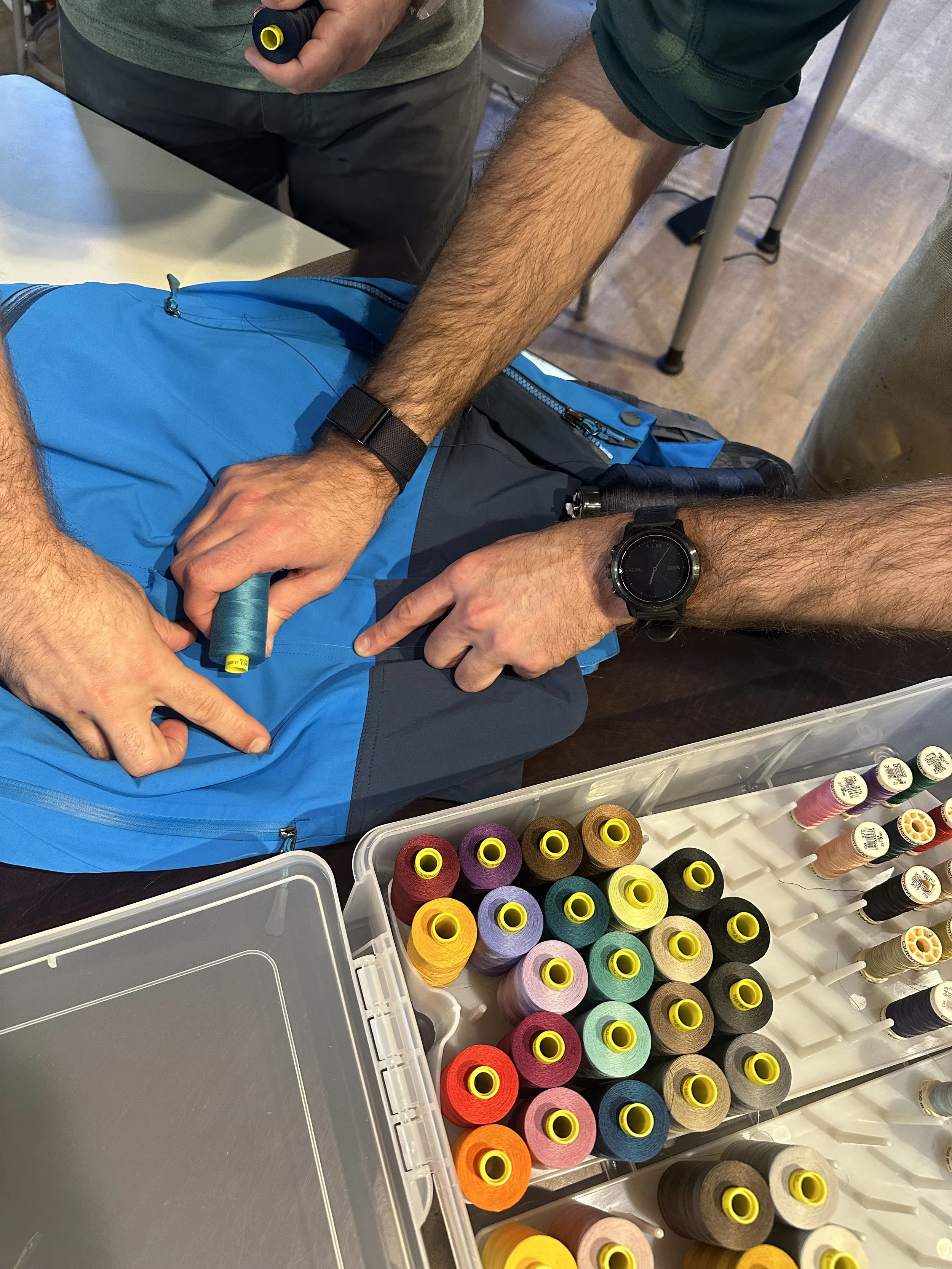 A man's hands hold a skein of blue thread next to a blue piece of clothing to determine if it is a match. An open box of thread is next to the fabric on the table.