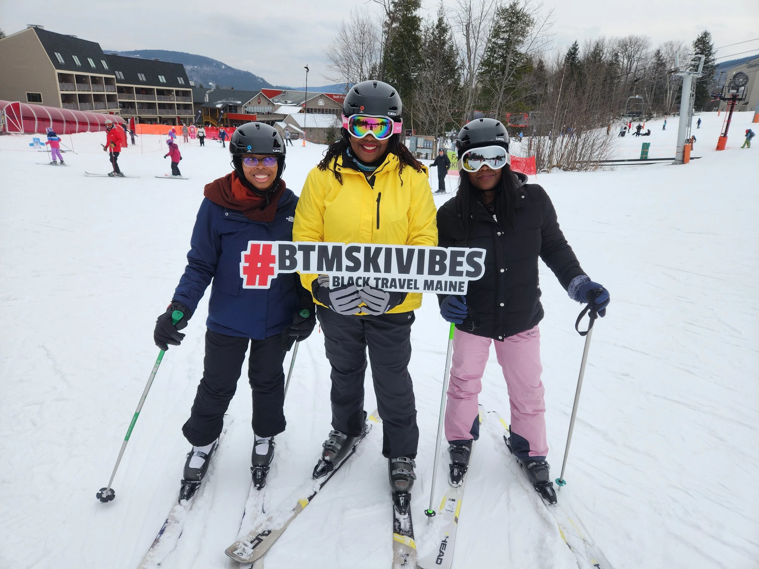 A group of a black boy, woman, and girl smile on a ski slope while holding a sign that says #btmskivibes