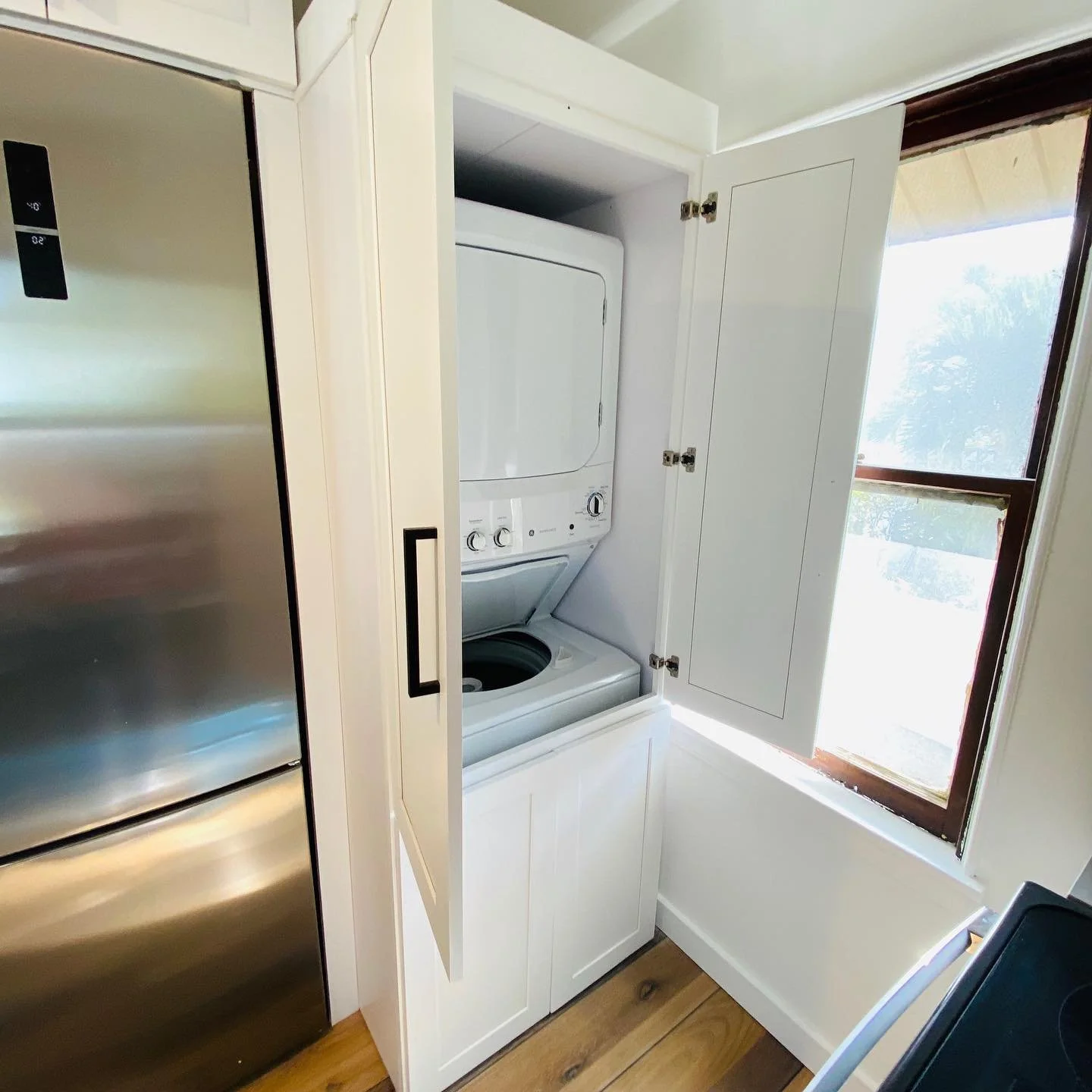 A stacked washer and dryer inside a white cabinet next to a window with a blind.