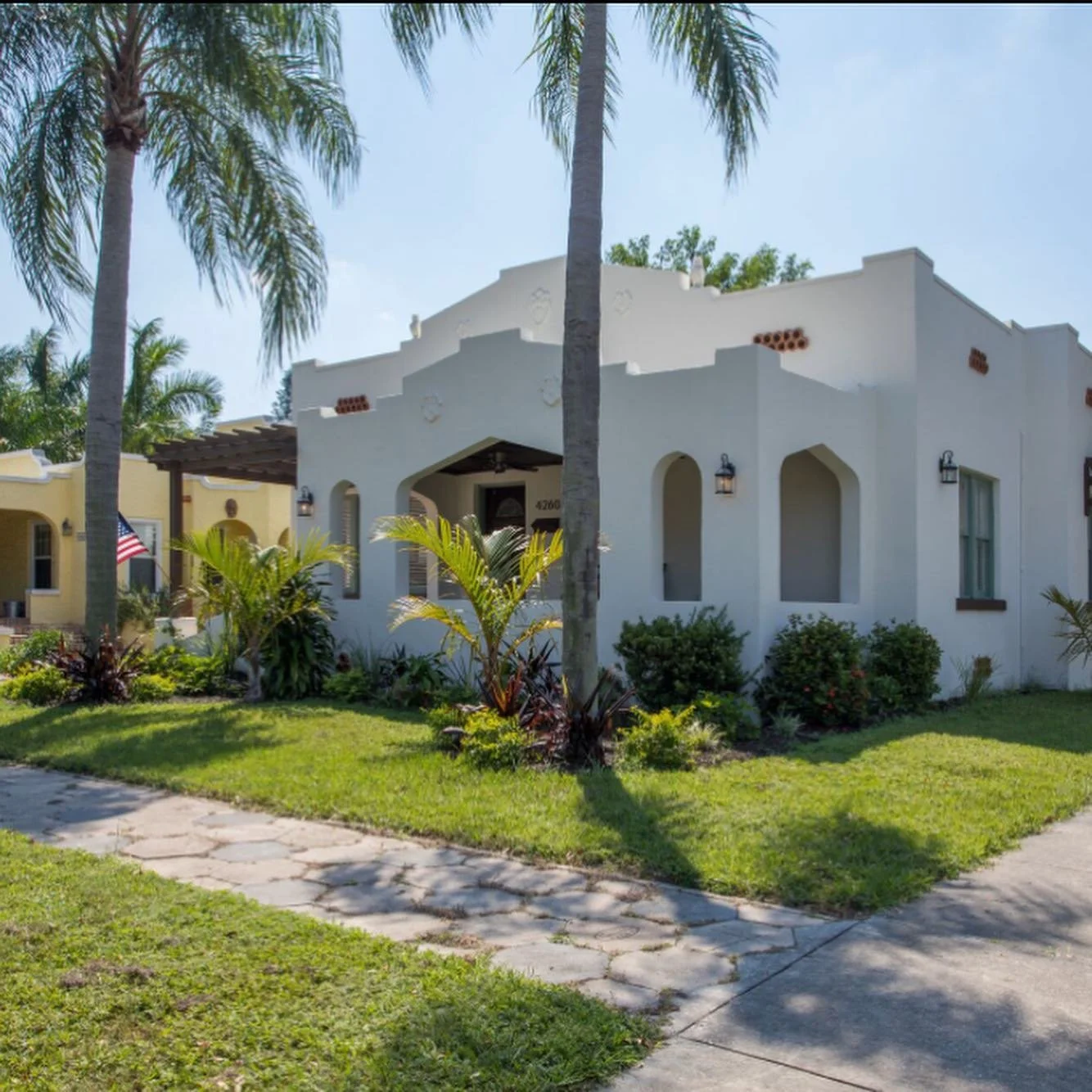 A white stucco house with arched windows and a flat roof, surrounded by palm trees and tropical plants, with a sidewalk and lush lawn in front, under a clear blue sky.