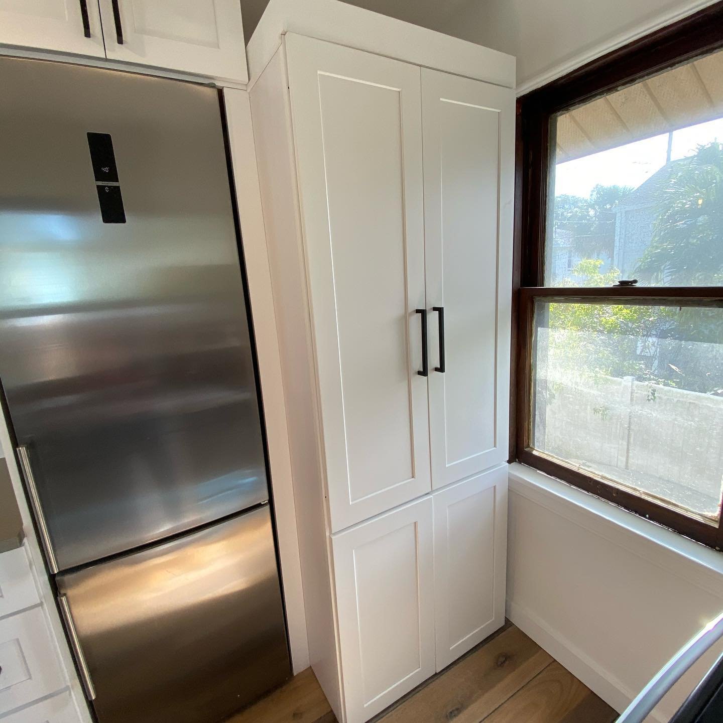 A kitchen corner with a stainless steel refrigerator on the left, a white cabinet with two doors and black handles in the middle, and a window with a brown frame on the right showing outdoor greenery and a neighboring house.