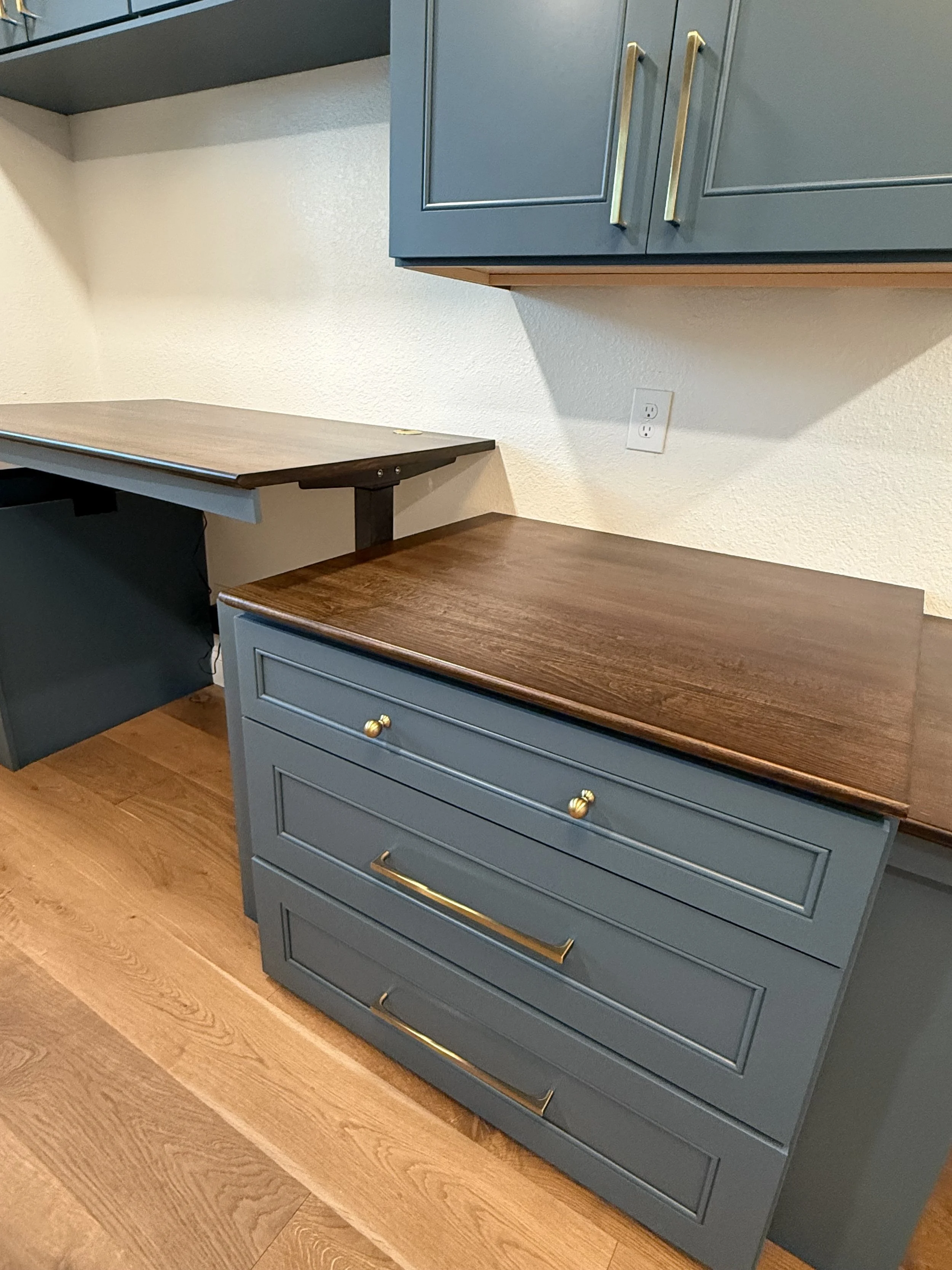 A kitchen with blue cabinets, gold handles, and drawers, with a brown wooden countertop. There is a beige wall with an electrical outlet and hardwood flooring.