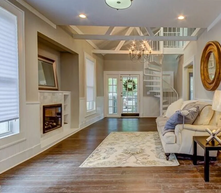 Living room with wooden floors, light-colored sofa, fireplace, large windows with blinds, a rug, and a staircase leading to an upper level.