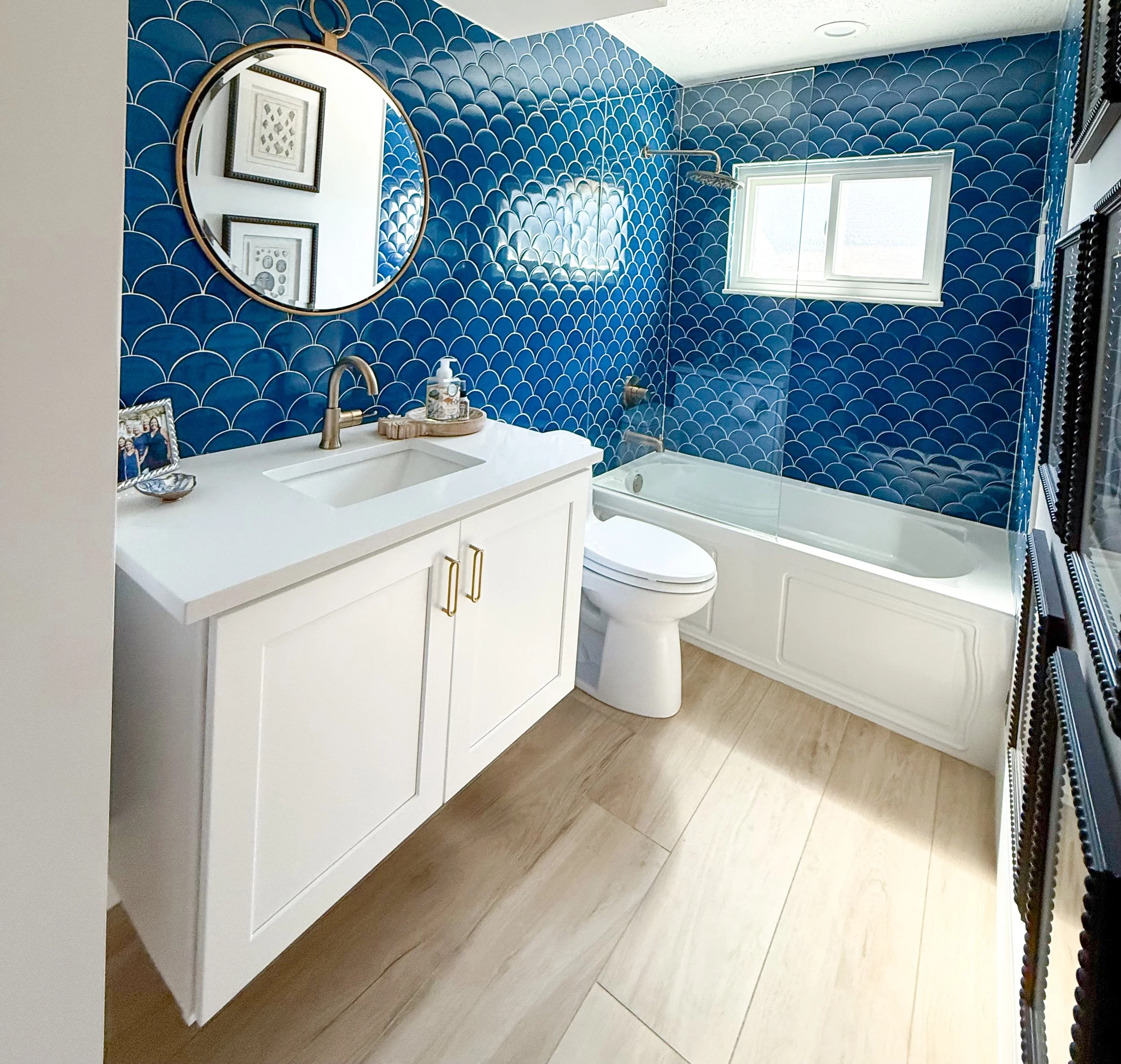 Bathroom with blue fishscale tile walls, a white vanity with gold handles, a round mirror, a toilet, a bathtub with a glass panel, a small window, and wood-look flooring.