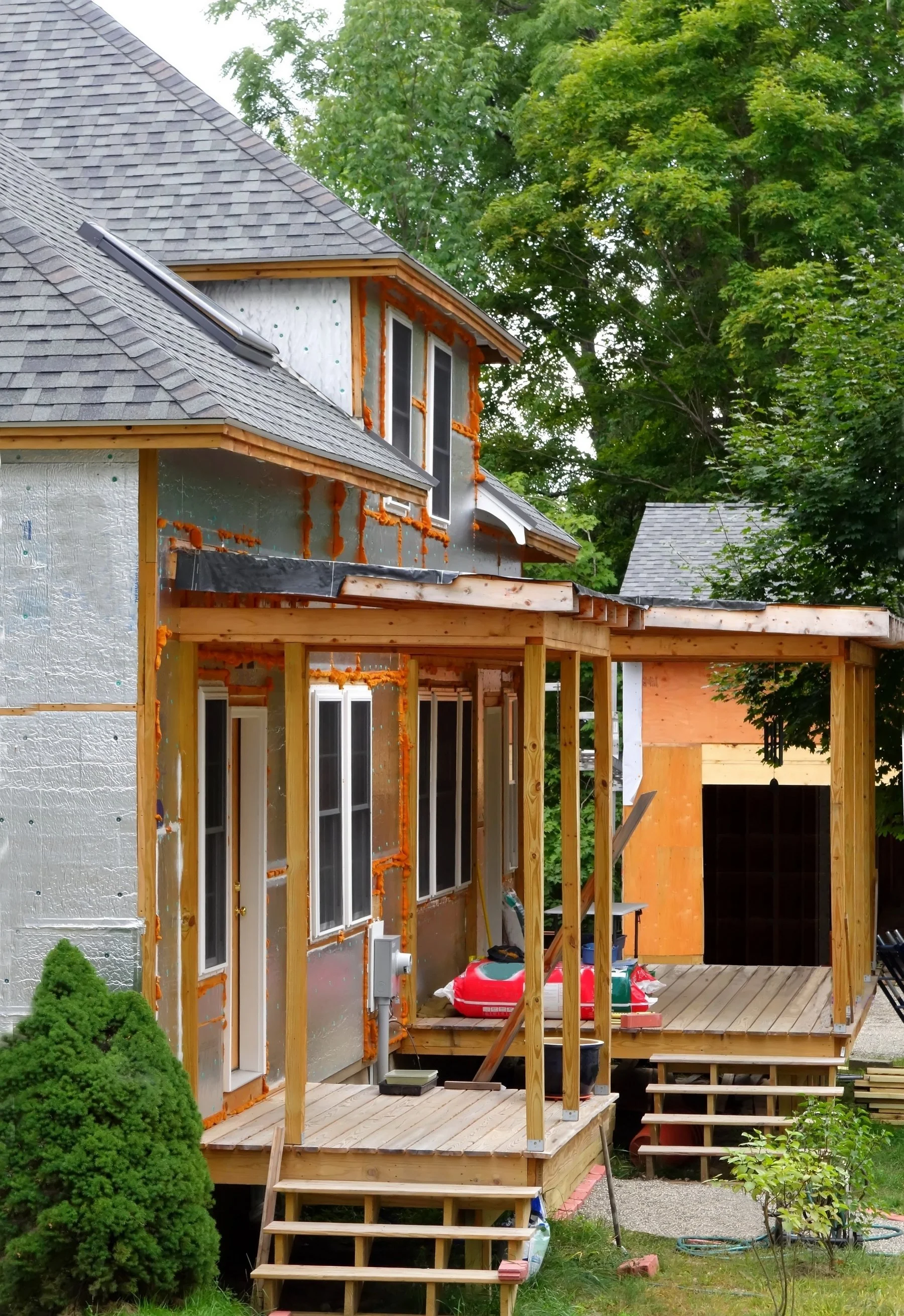 A house under construction with wooden framing and insulation, surrounded by green trees, with a partially built porch and construction tools.