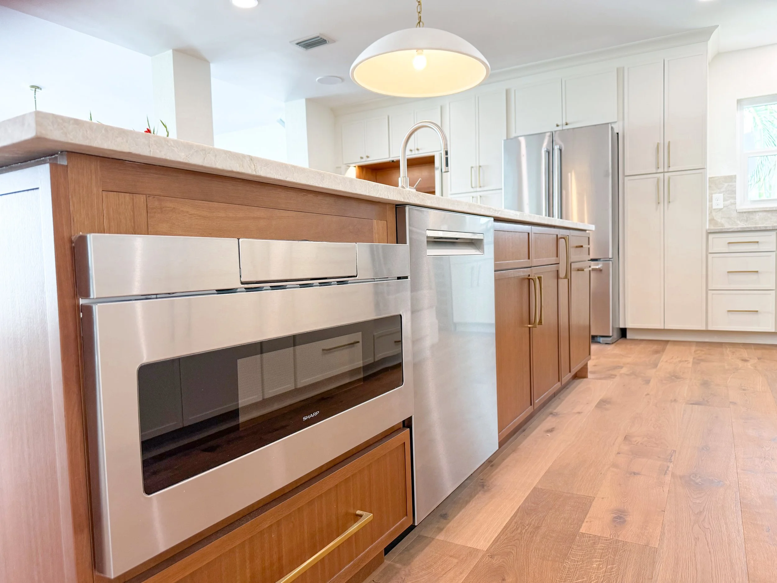 Modern kitchen with wooden and white cabinetry, stainless steel appliances including a microwave and refrigerator, light-colored countertops, and hardwood flooring.