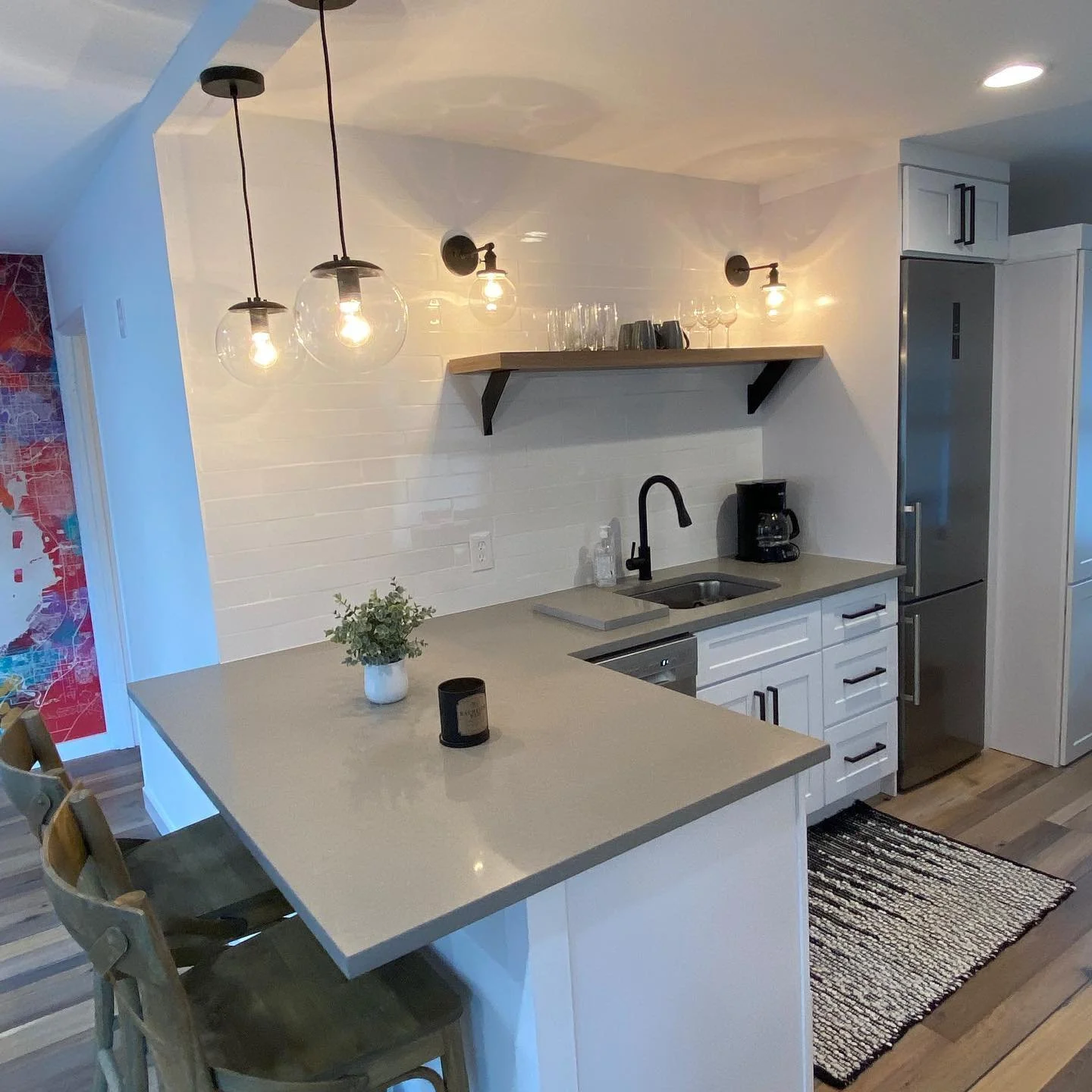Modern kitchen with white cabinets, a beige countertop, and a stainless steel refrigerator. There are three hanging pendant lights with glass globes and Edison bulbs. A black faucet is above the sink, and a coffee maker is on the counter. A wooden sh