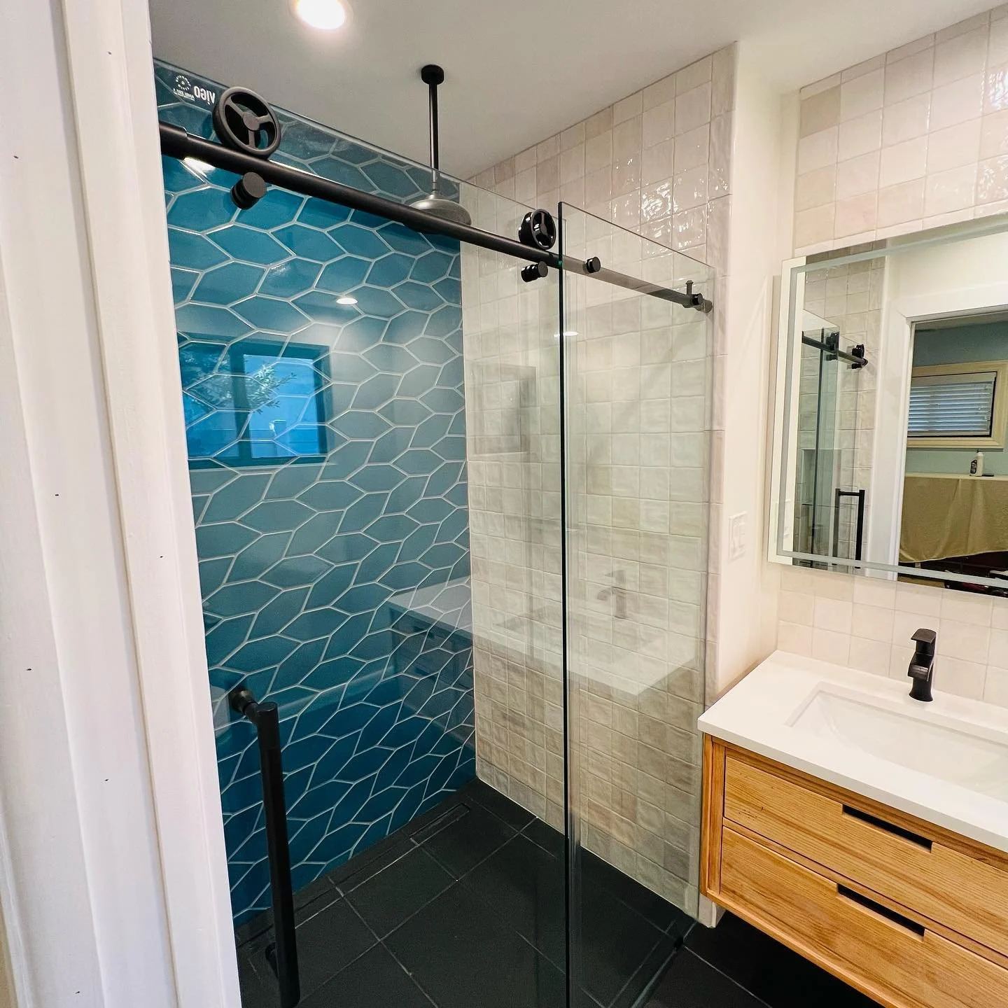Modern bathroom with a walk-in shower featuring blue hexagonal tiles, a black sliding barn door track, and a wooden vanity with a white sink and black faucet, a large mirror, and cream-colored tiled walls.