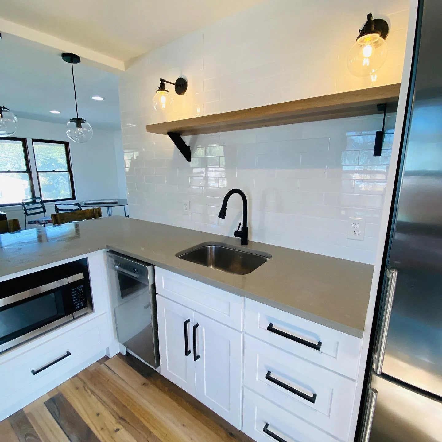 Modern kitchen with white cabinets, beige countertop, black faucet, open wooden shelf, stainless steel microwave, small fridge, and a white backsplash with subway tiles.