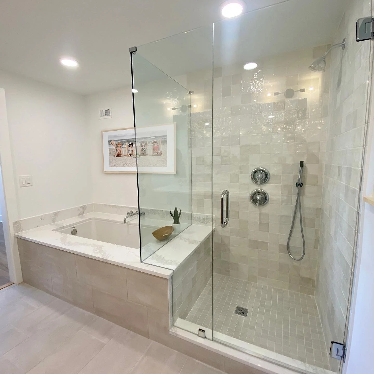 A modern bathroom with a glass-enclosed shower area and a bathtub, decorated with neutral-toned tiles and minimalist accessories, including a framed beach scene on the wall.