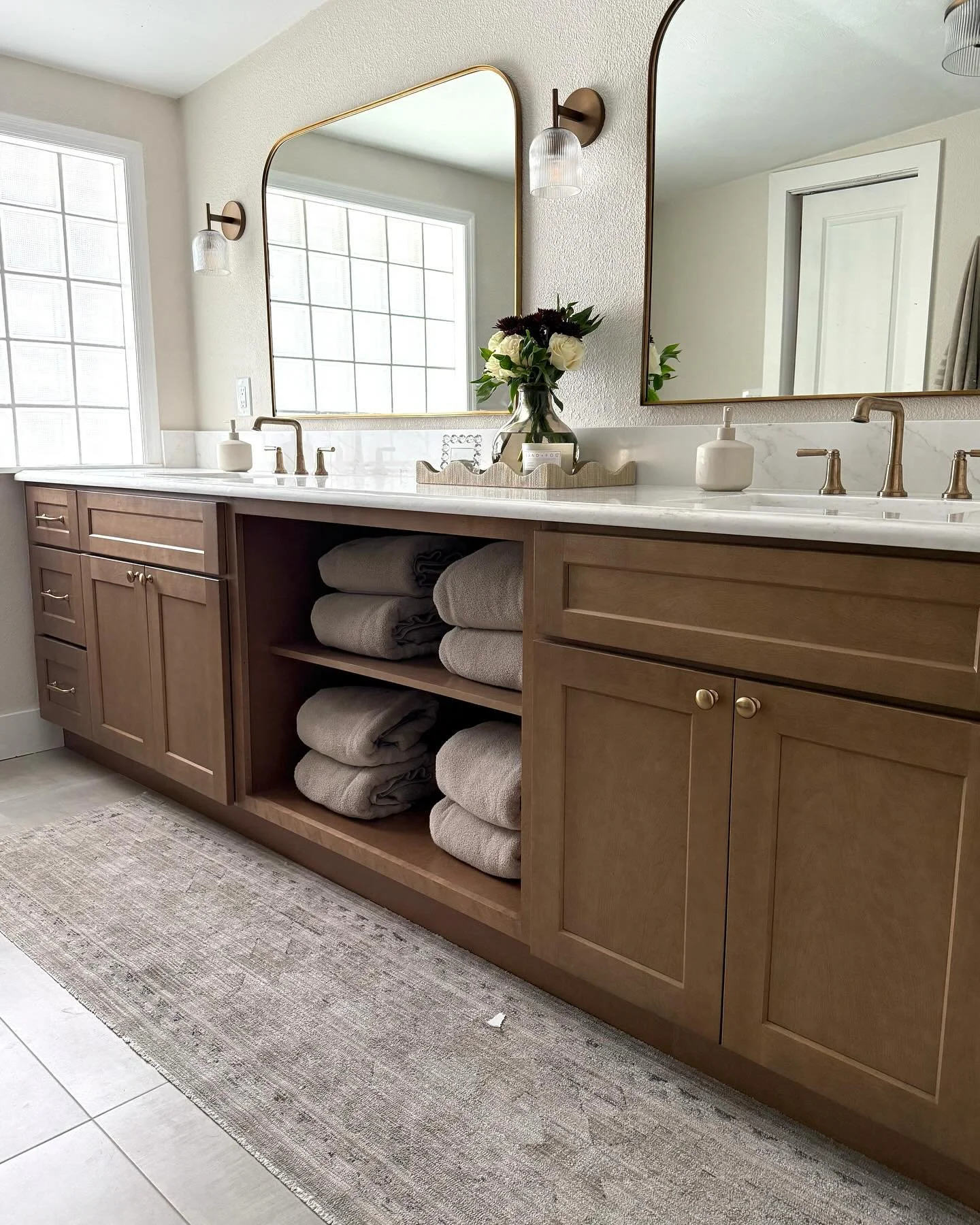 Bathroom vanity with beige wooden cabinets and open shelves holding folded towels, granite countertop with two sinks, large mirrors, wall-mounted lamps, a vase with flowers, and a window with frosted glass blocks.