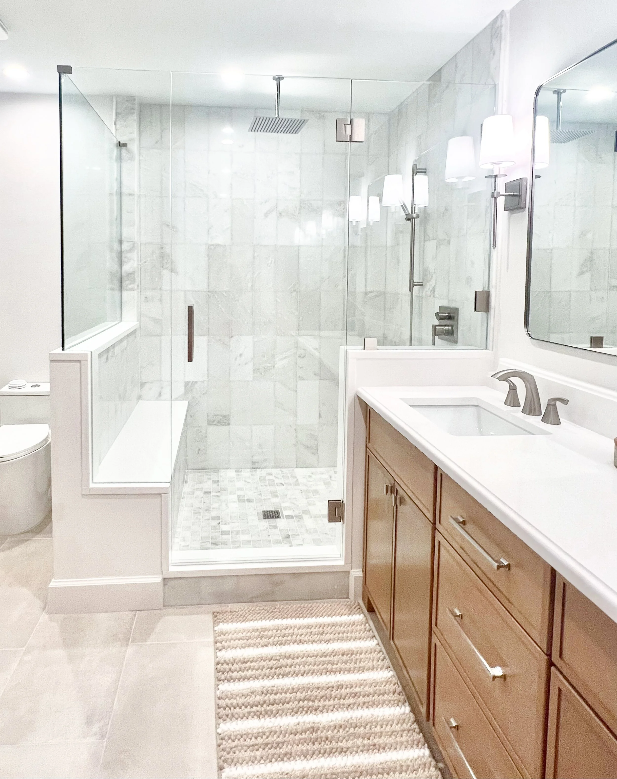 Modern bathroom with glass-enclosed shower, white marble tiles, wooden vanity with a white sink, large mirror, and striped rug on beige tile floor.