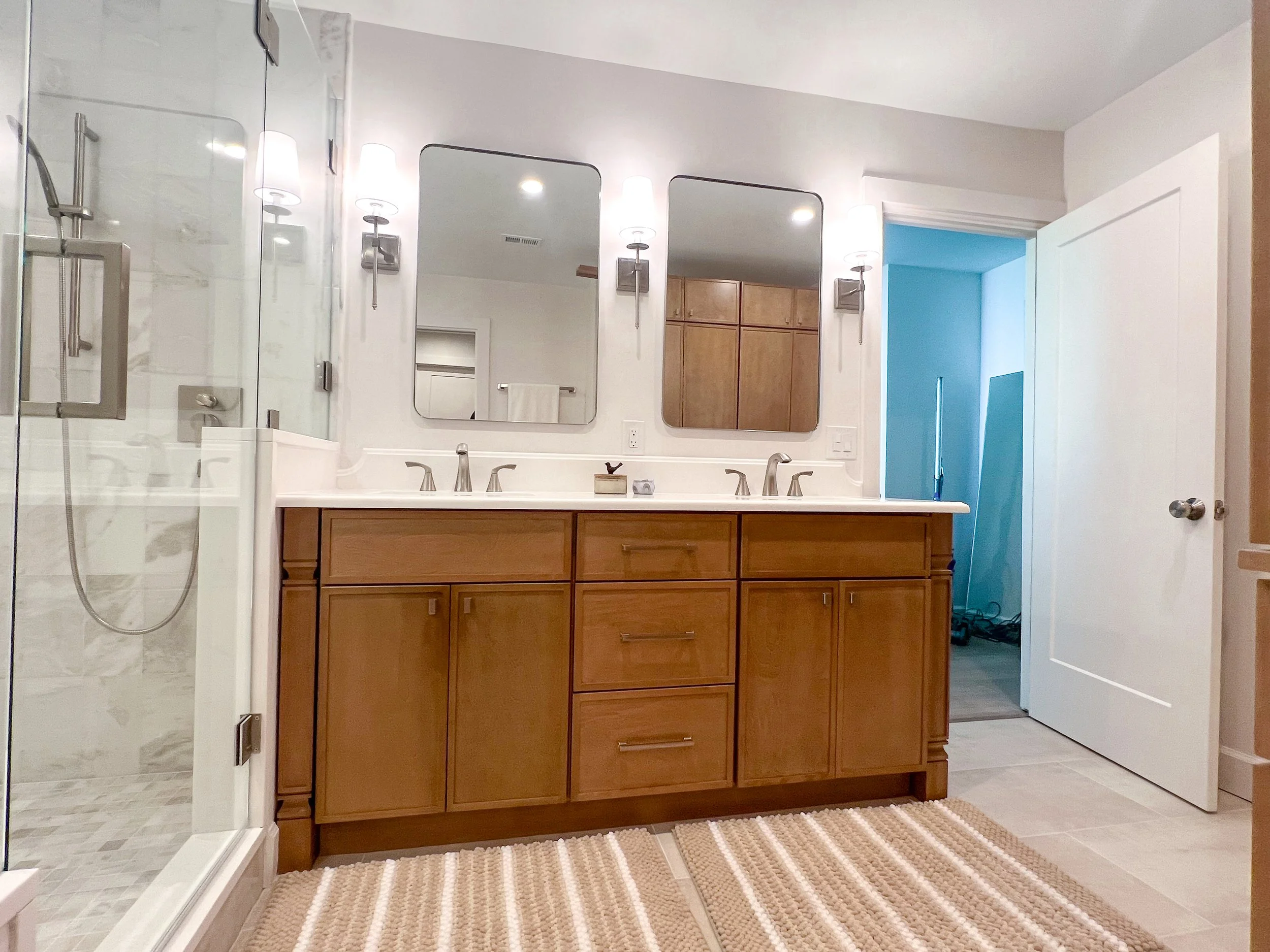 A bathroom with a double sink vanity with wood cabinets, two mirrors, and wall-mounted lights. To the left, there is a glass-enclosed shower. The floor has beige tiles, and a striped beige and white rug. An open door leads to a room with blue walls a