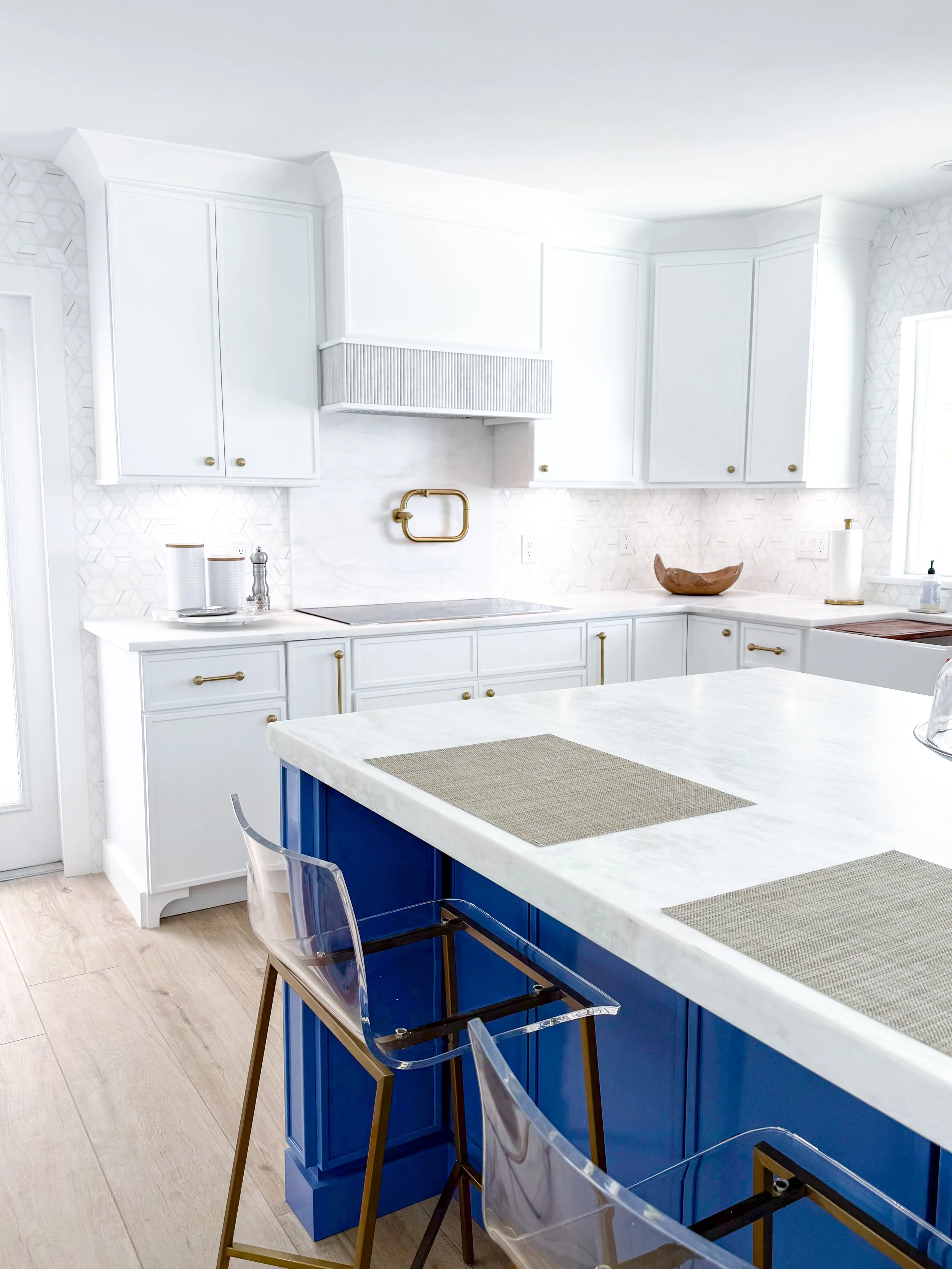 Modern kitchen with white cabinets, a blue island, and clear acrylic chairs. White countertops and natural light from a nearby window.